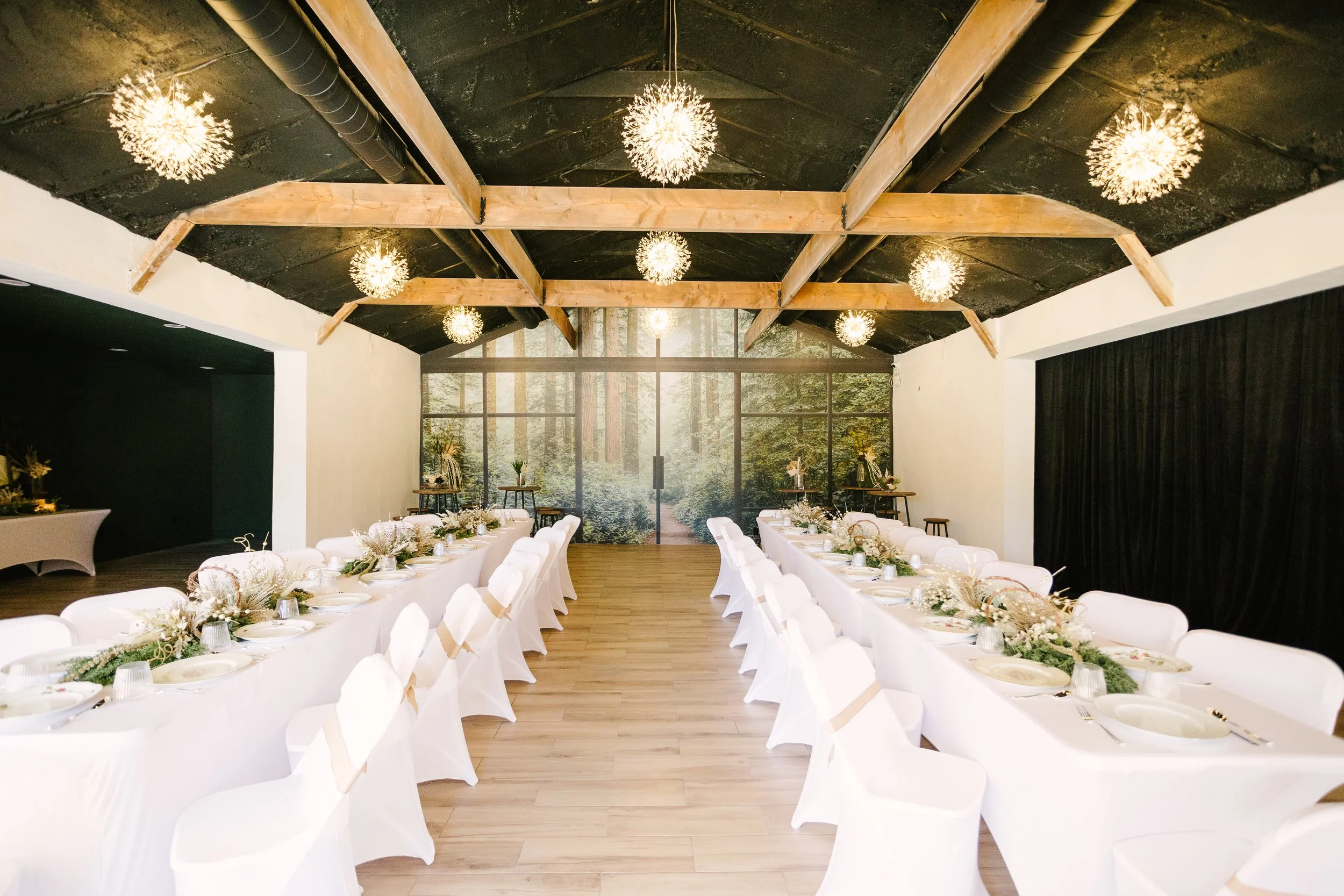 Elegant event space with long white banquet tables, white chairs, floral arrangements, and a large forest mural on the back wall.