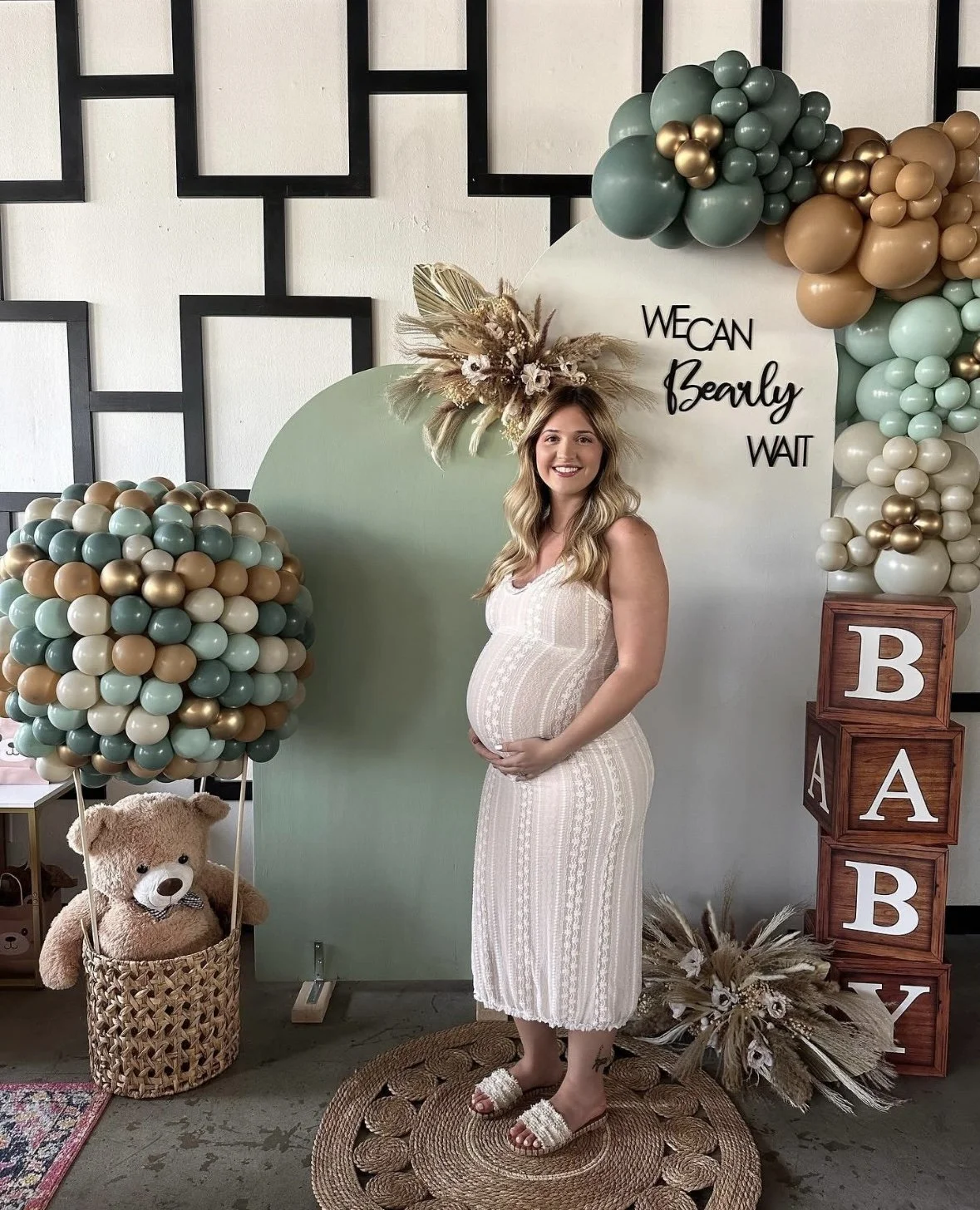 Pregnant woman with long blonde hair in a white dress standing on a woven rug, smiling and holding her belly, surrounded by balloons, a teddy bear in a basket, flowers, and decorative blocks spelling 'BABY' at a baby shower or maternity event.