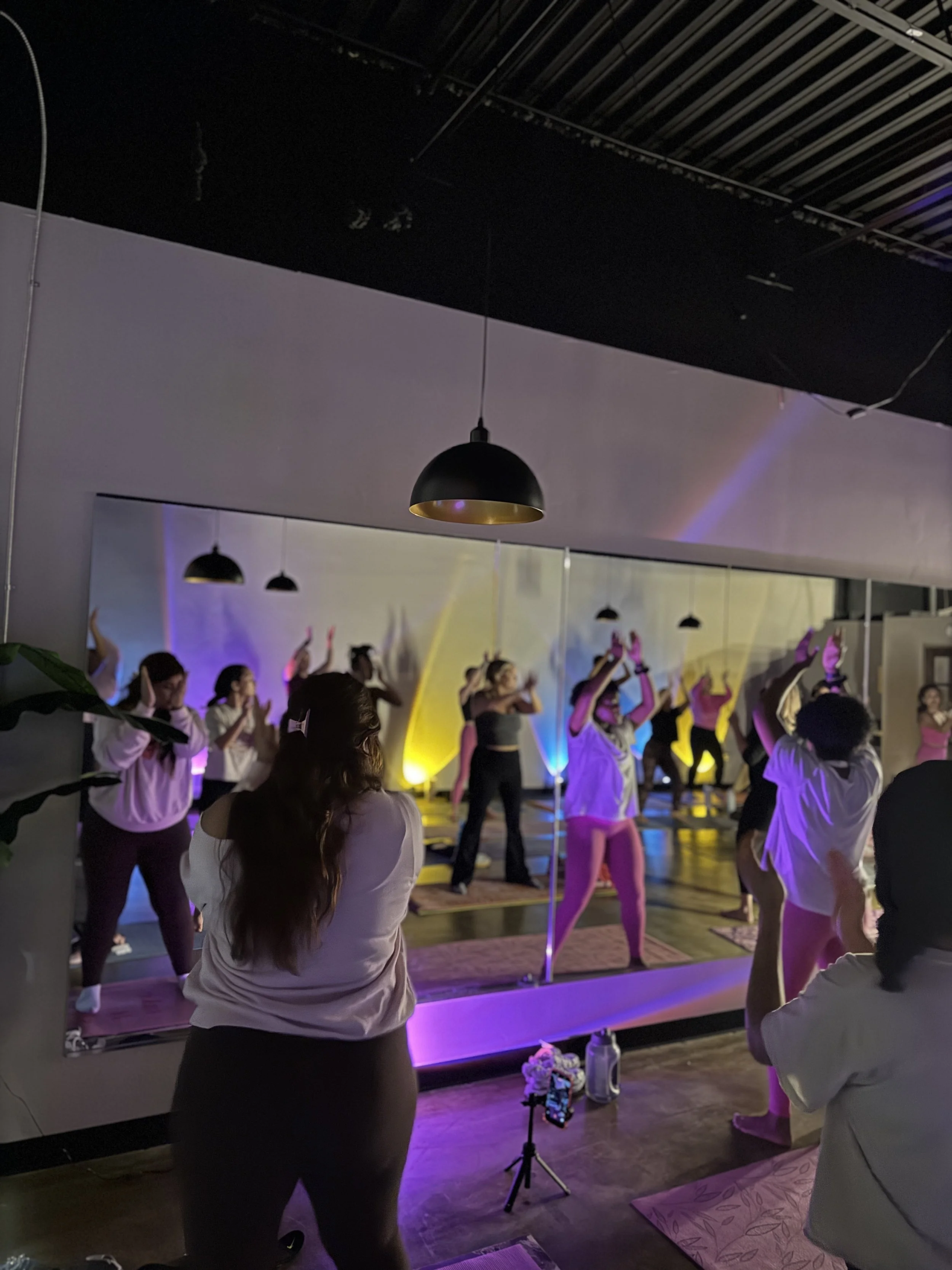A group of people practicing yoga in a fitness studio with colorful lighting, viewed through a mirror on the wall.