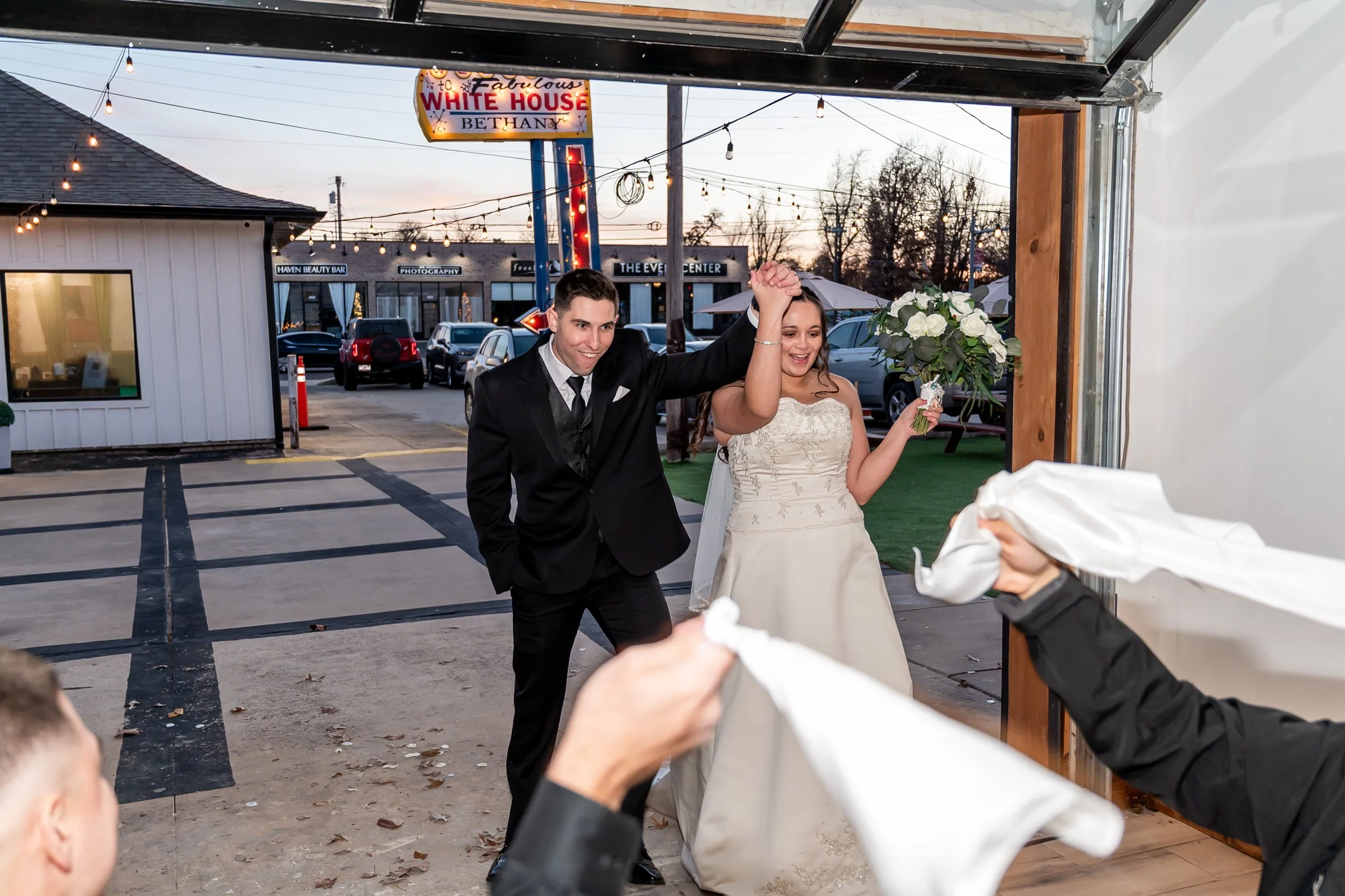 A newlywed couple is dancing at their wedding reception, smiling and holding hands with the groom wearing a black tuxedo and the bride in a white wedding dress holding a bouquet of white flowers. Guests are seen throwing napkins or cloths at them.