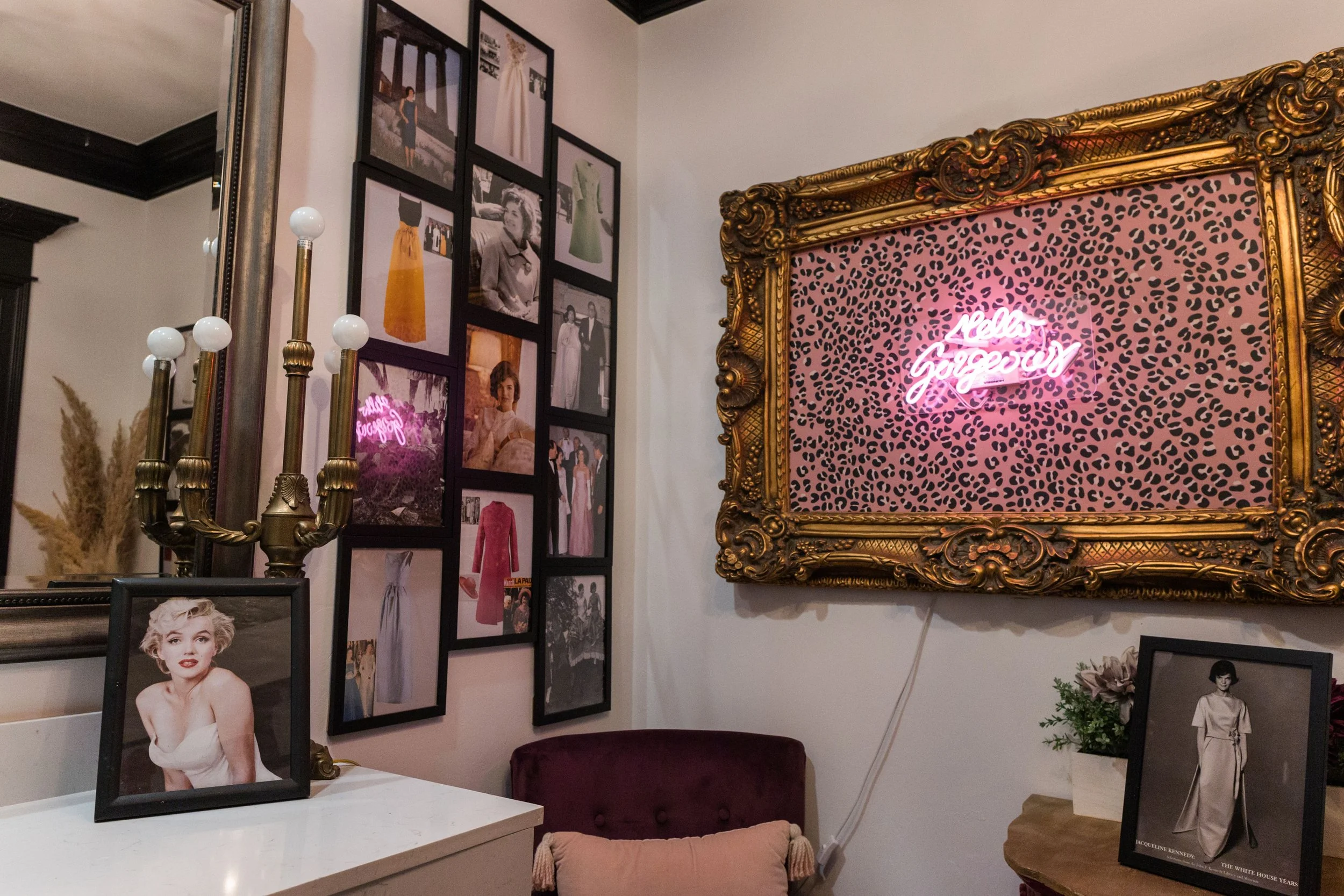 A corner of a room decorated with framed photos, a vintage mirror, a gilded ornate frame with neon pink and white text saying "Nallas Gingen," a small white table with a framed photo of Marilyn Monroe, a purple chair with a beige cushion, a bunch of 