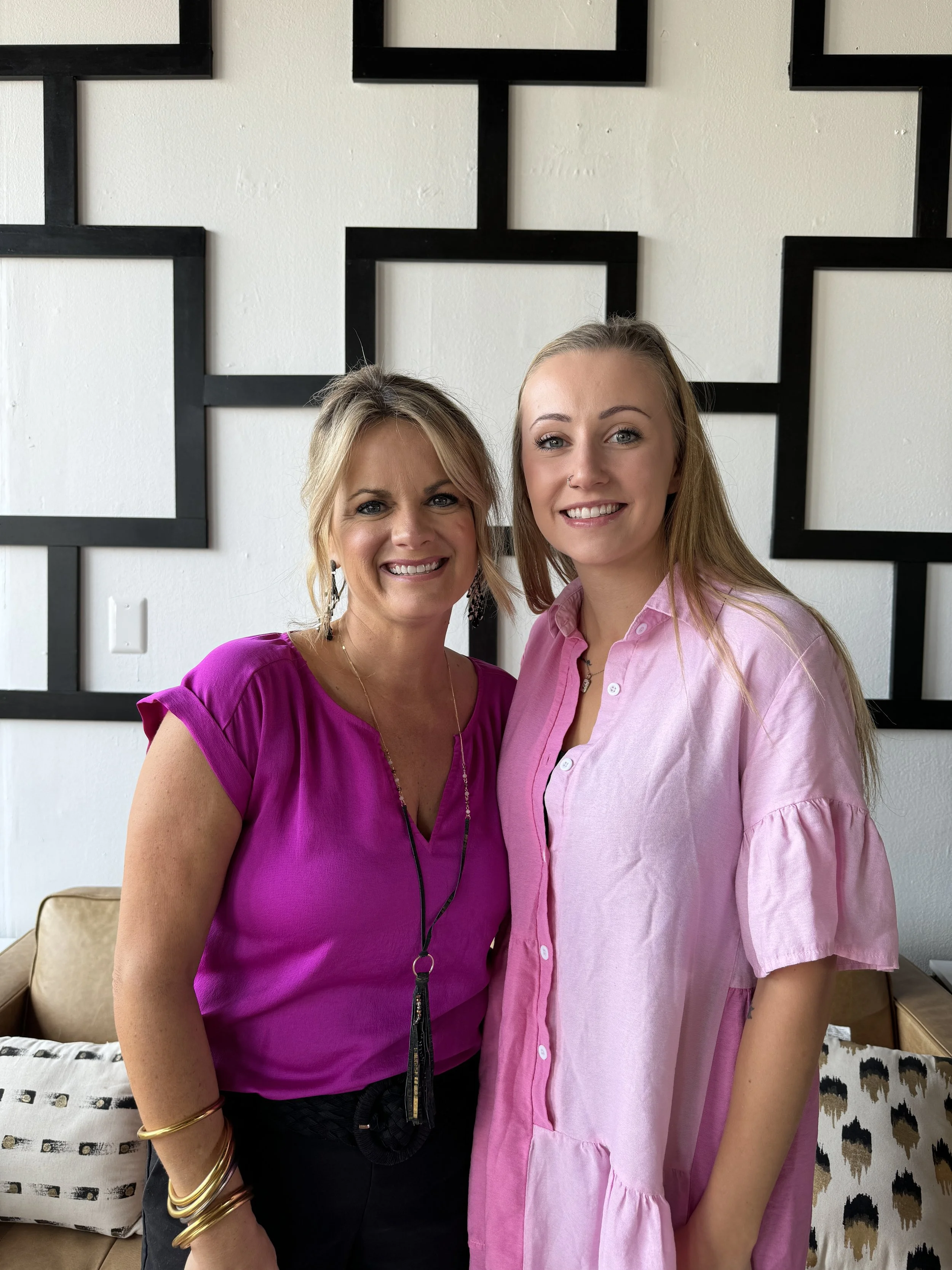 Two women standing together, smiling, in front of a modern black and white geometric wall art. The woman on the left has blonde hair, wearing a bright pink top with gold jewelry. The woman on the right has light brown hair, wearing a light pink butto