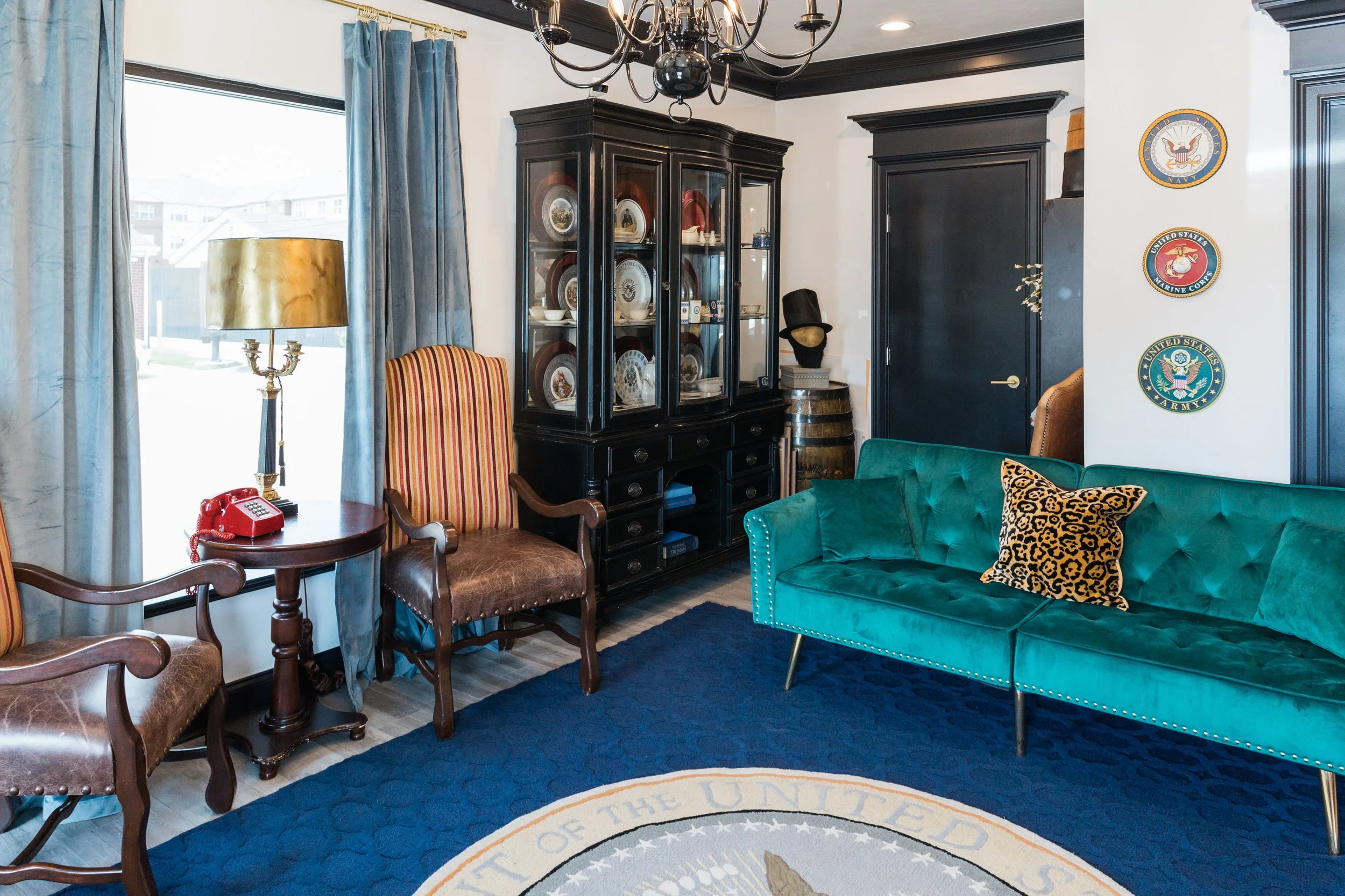 Living room with teal velvet sofa, leopard print pillow, black china cabinet with dishes, wooden chairs, gold table lamp, red vintage telephone, decorative wall plates with military insignia, blue curtains, dark painted wood trim, chandelier, window 