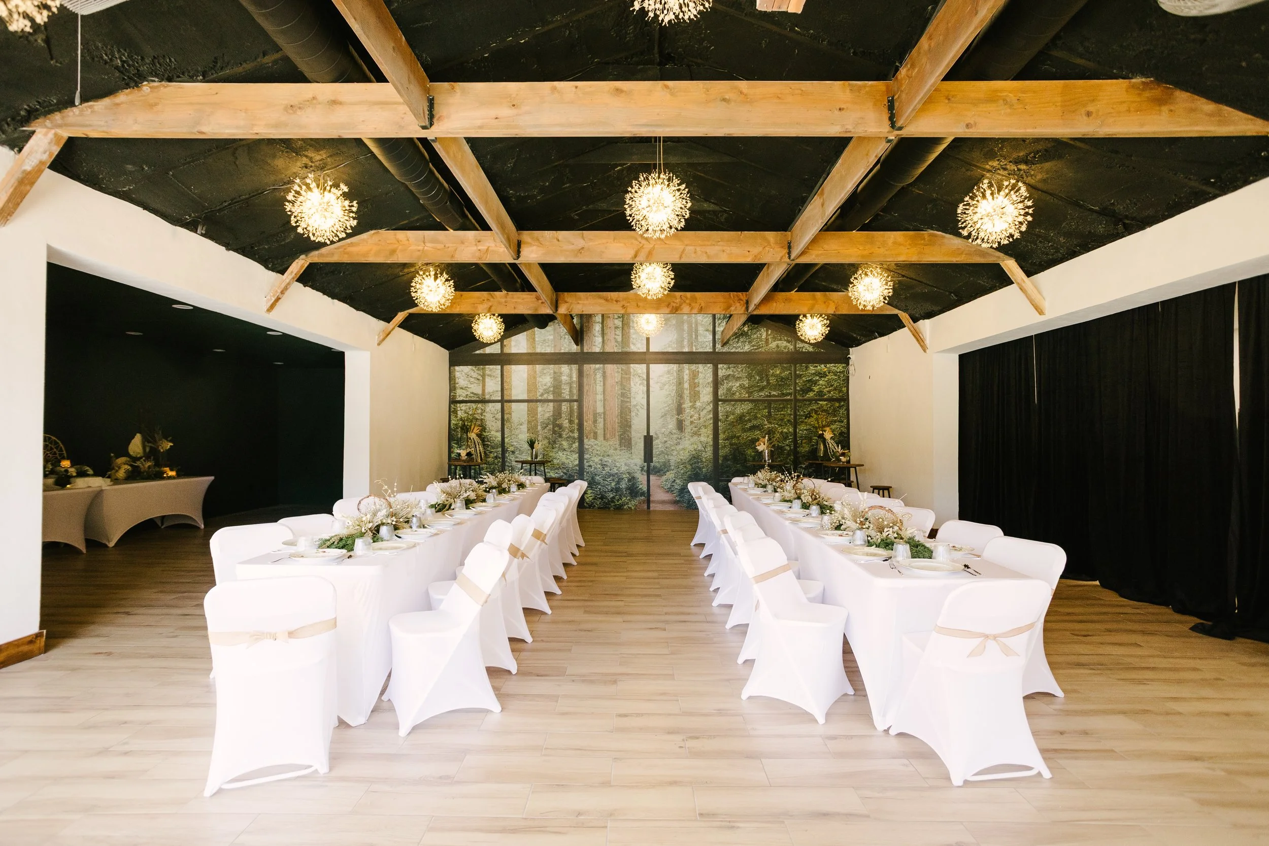 Elegant indoor wedding reception with long white-draped tables, white chairs with beige ribbons, floral centerpieces, and a large window showing a forest view.