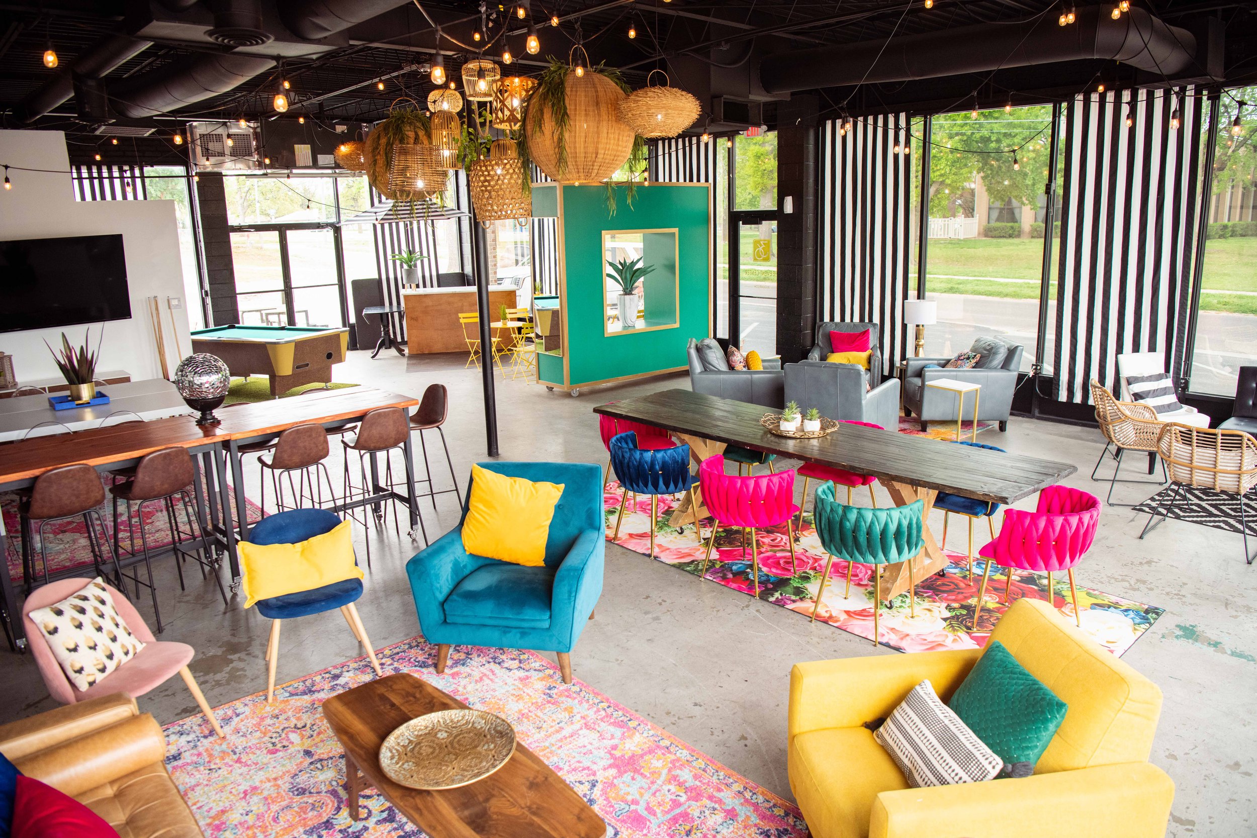 A brightly decorated lounge or community space with colorful chairs, sofas, and tables. There are hanging lights, plants, a pool table, and large windows with striped blinds providing natural light.