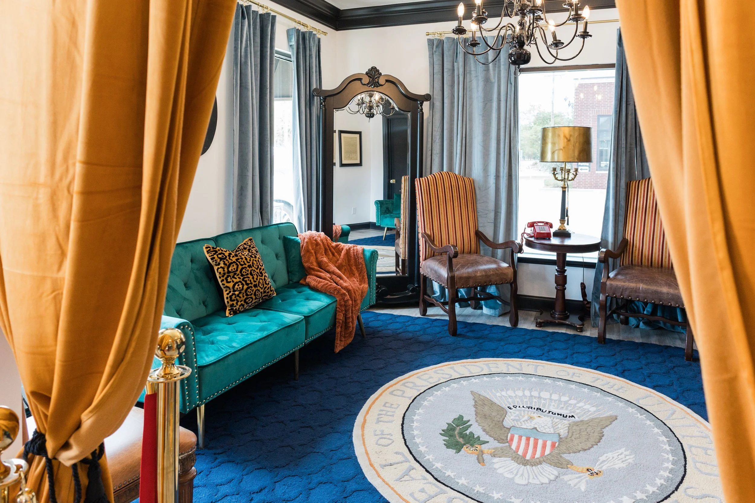 A luxurious room decorated with a teal velvet sofa, an orange throw blanket, and a leopard print pillow. The room features a navy blue carpet with the presidential seal of the United States in the center, large windows with blue curtains, a wooden mi