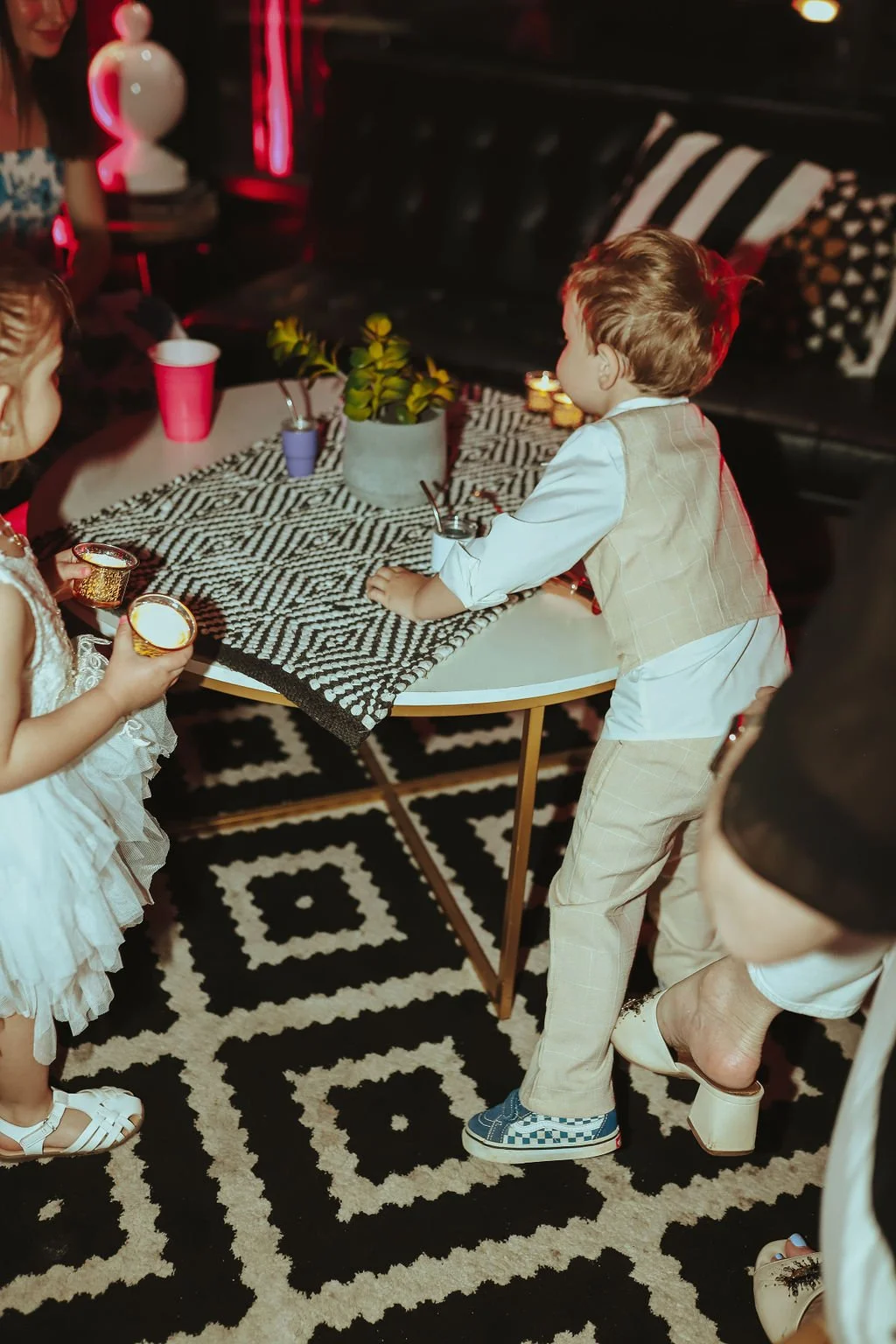 Children at a party around a table with candles, drinks, and a potted plant, in a room with black and white decor.