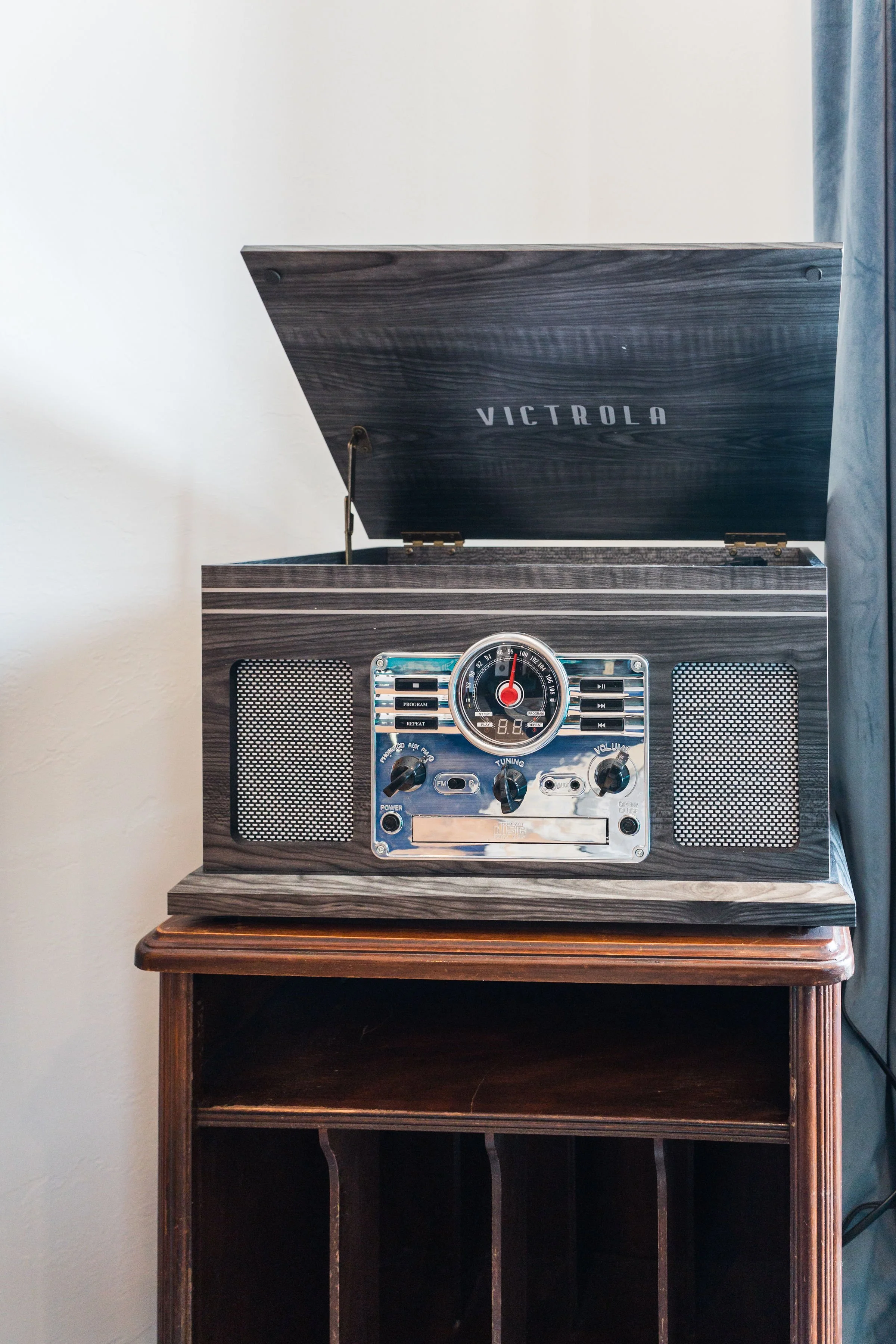 Vintage Victrola record player with dark wood case and silver control panel, on a wooden stand, with blue curtains on the side.