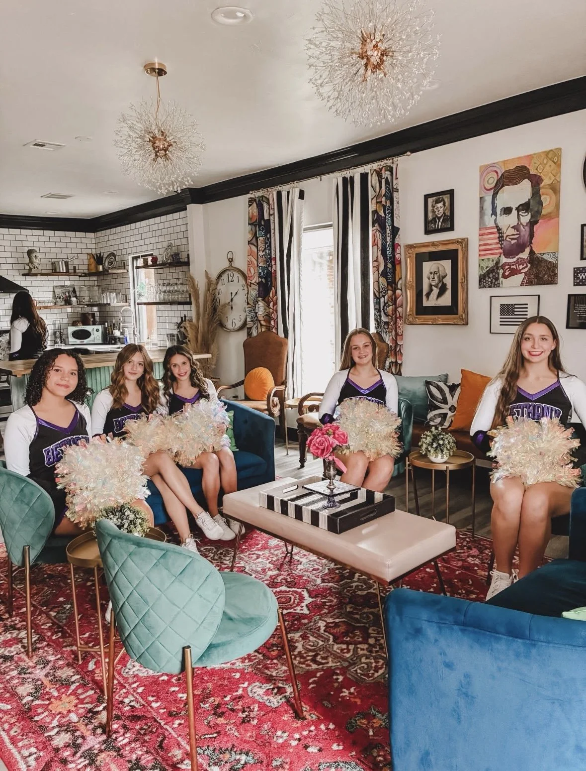 Five cheerleaders in matching uniforms sitting on sofas in a living room with colorful décor, holding pom-poms and smiling.