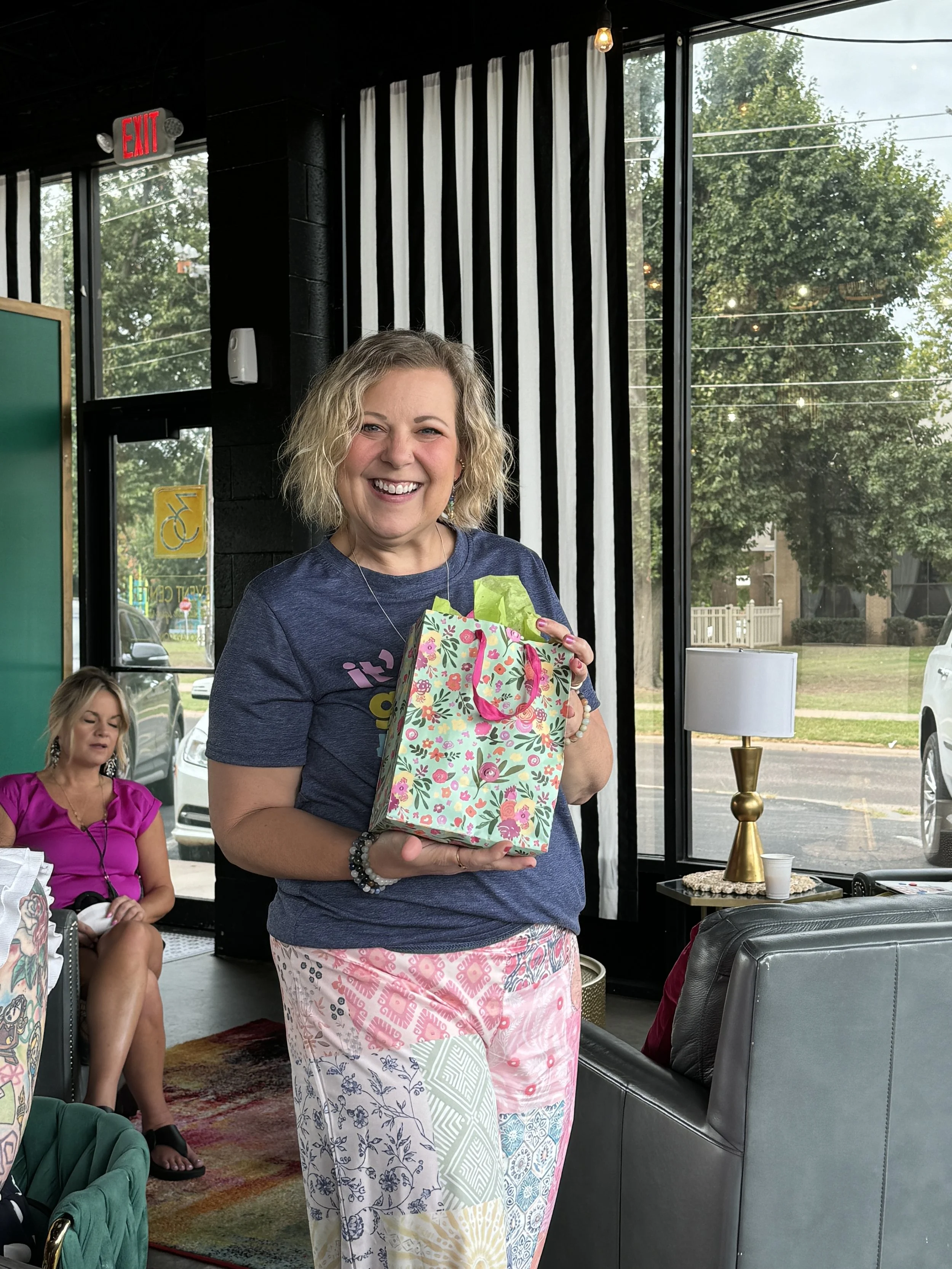 A smiling woman with curly blonde hair holding a colorful gift bag, inside a restaurant with large windows, black and white striped curtains, and a group of women sitting nearby.