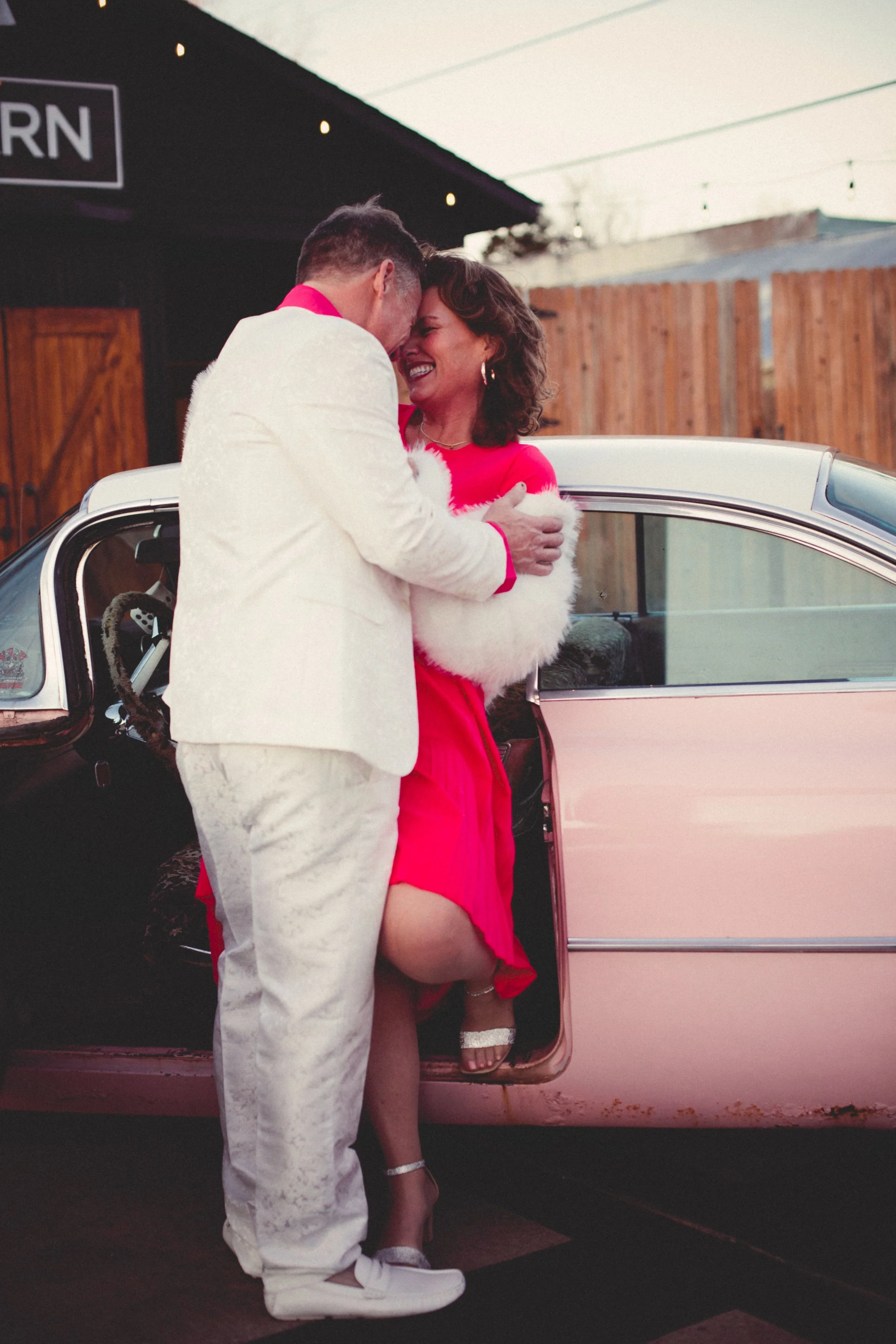A couple dressed in festive holiday attire sharing a joyful moment outside a vintage pink car.