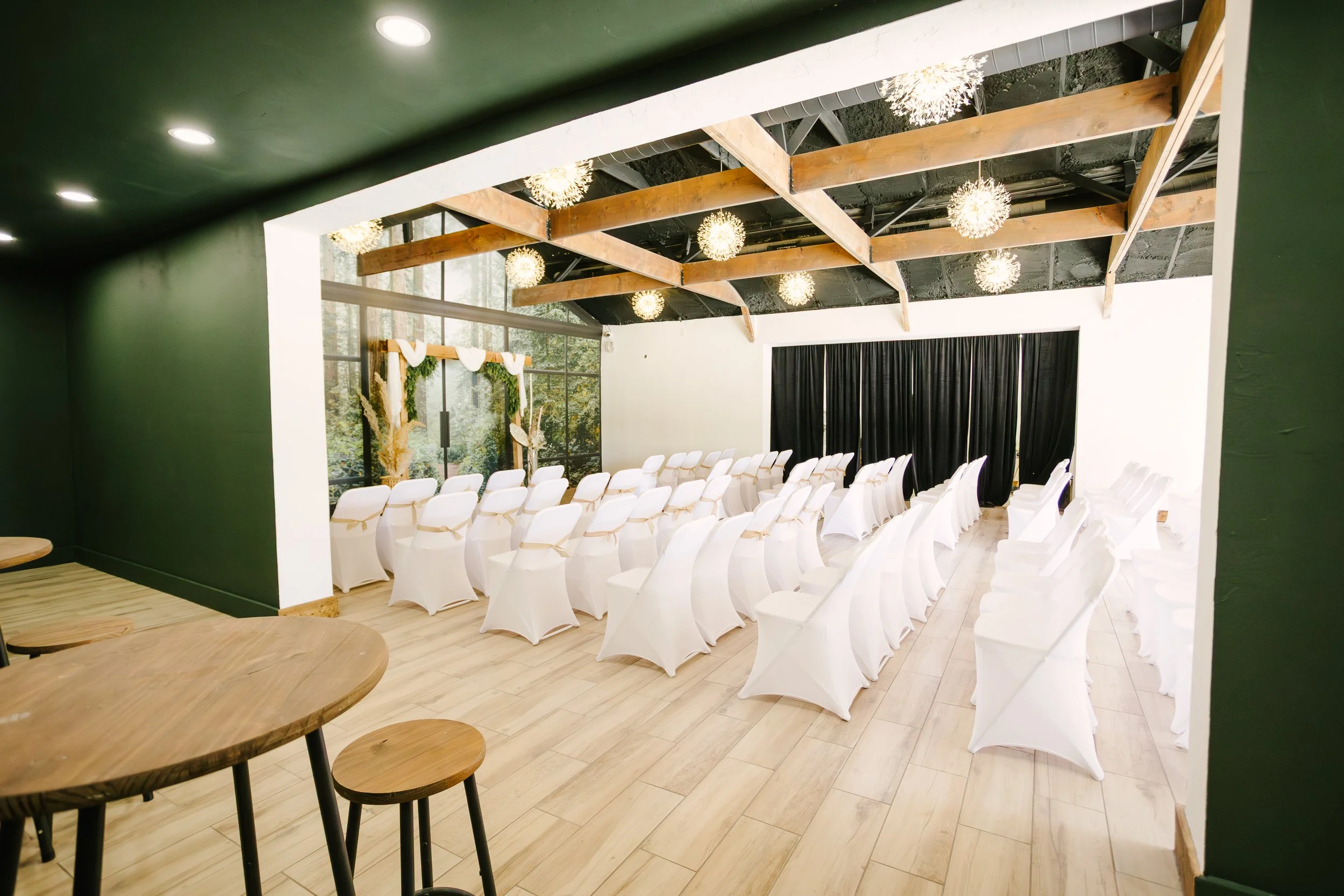 Wedding ceremony setup with rows of white chairs covered in white fabric and tied with beige ribbons, a decorative arch with greenery and white drapery at the front, black curtains behind the arch, large windows with trees visible outside, wooden bea