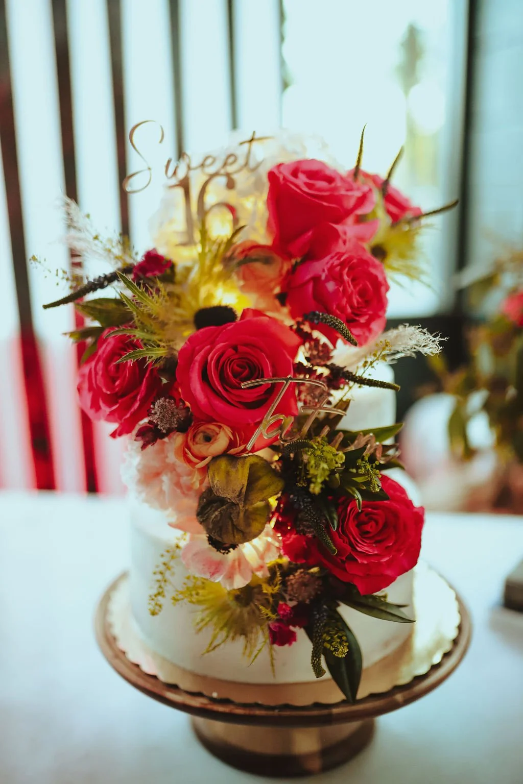 A birthday cake decorated with pink and red roses, greenery, and a topper that says 'Sweet 16'. The cake is on a stand set on a table.