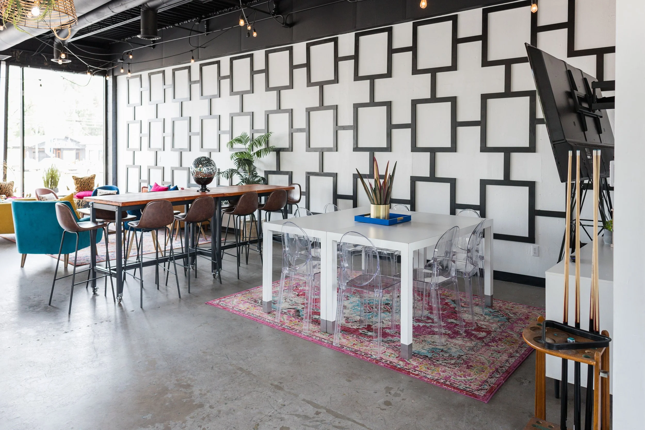 Interior of a modern room with a long wooden table, colorful chairs, a white square table with transparent chairs, a patterned rug, and wall decoration of square frames, with large windows and ceiling string lights.