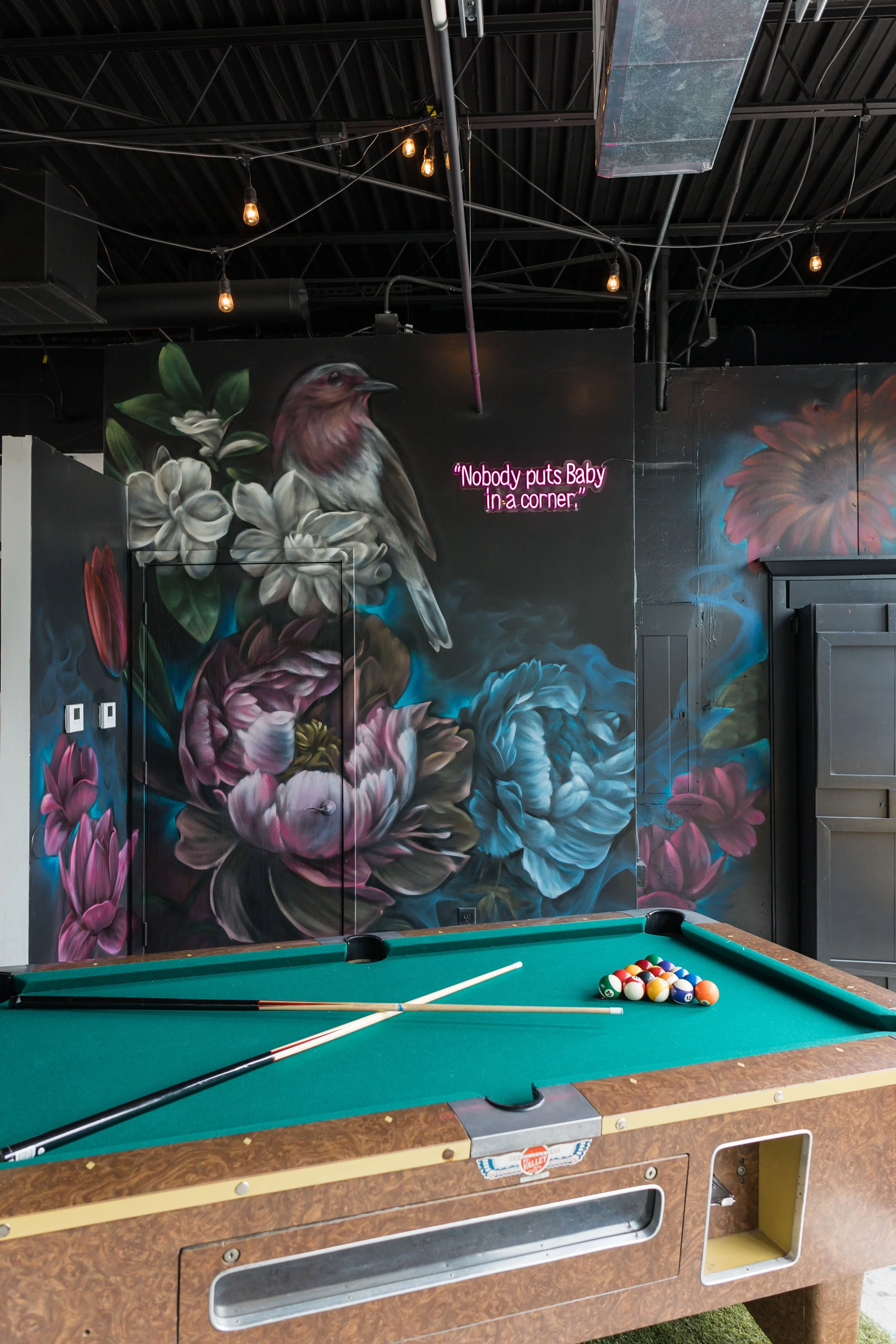 Interior of a room with a pool table and colorful floral mural on the wall, featuring a bird and the phrase "Nobody puts Baby in a corner" in neon pink letters.