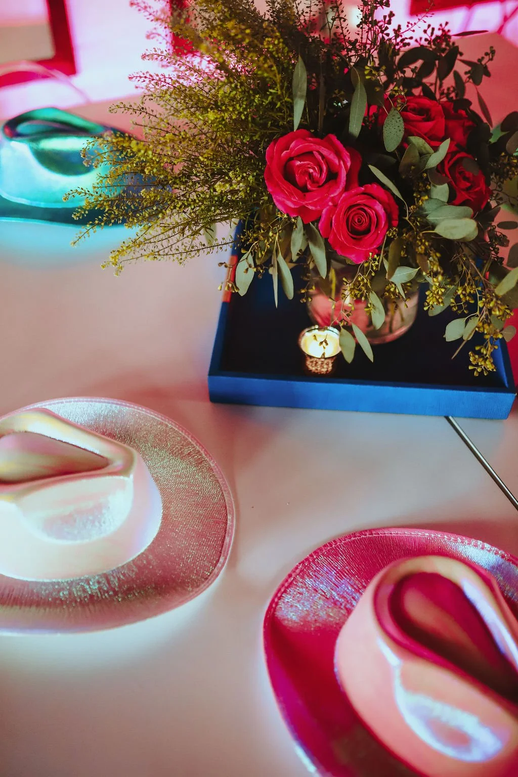 A table with a flower arrangement of red roses and greenery in a blue box, a small decorative candle, and two plates of desserts with pink and gold accents in a pink-lit setting.