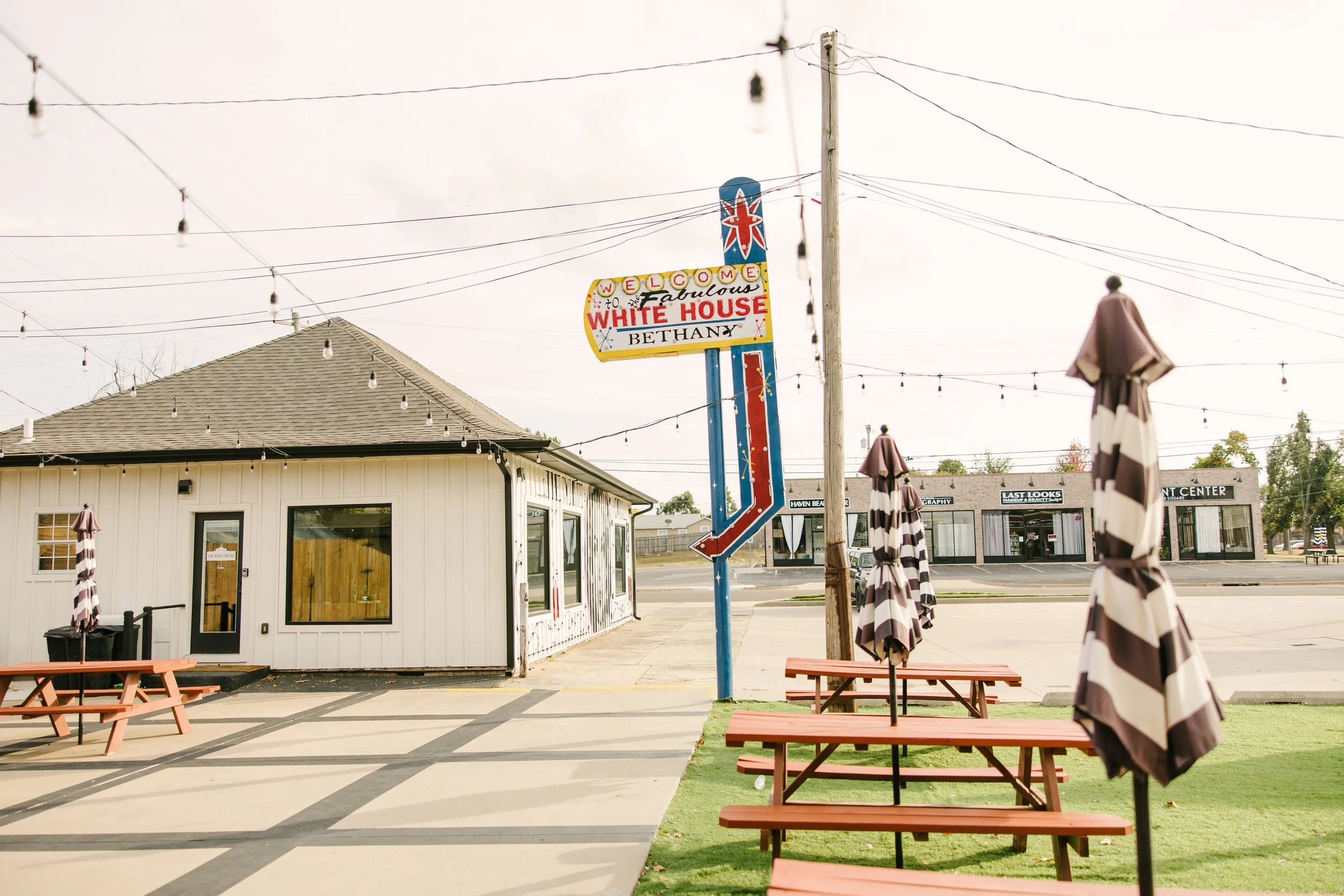 Empty outdoor patio with striped umbrellas and wooden picnic tables, a small white building with windows and a door, and a colorful sign that reads 'Welcome to Fabulous White House Bethany'. String lights are hanging above the patio, and across the s