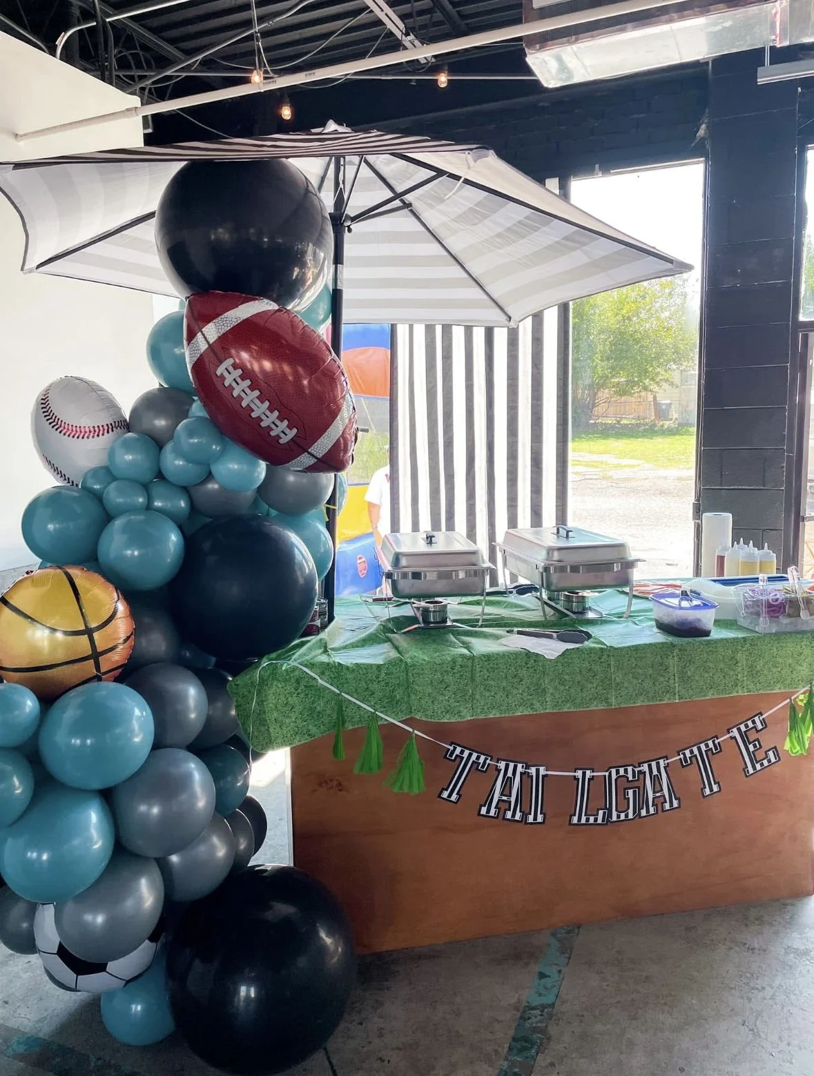 Party table decorated with balloons shaped like various sports balls, including football, baseball, basketball, soccer, and tennis, with a 'Tackle Tag' banner, set up under a large umbrella with food trays and condiments.