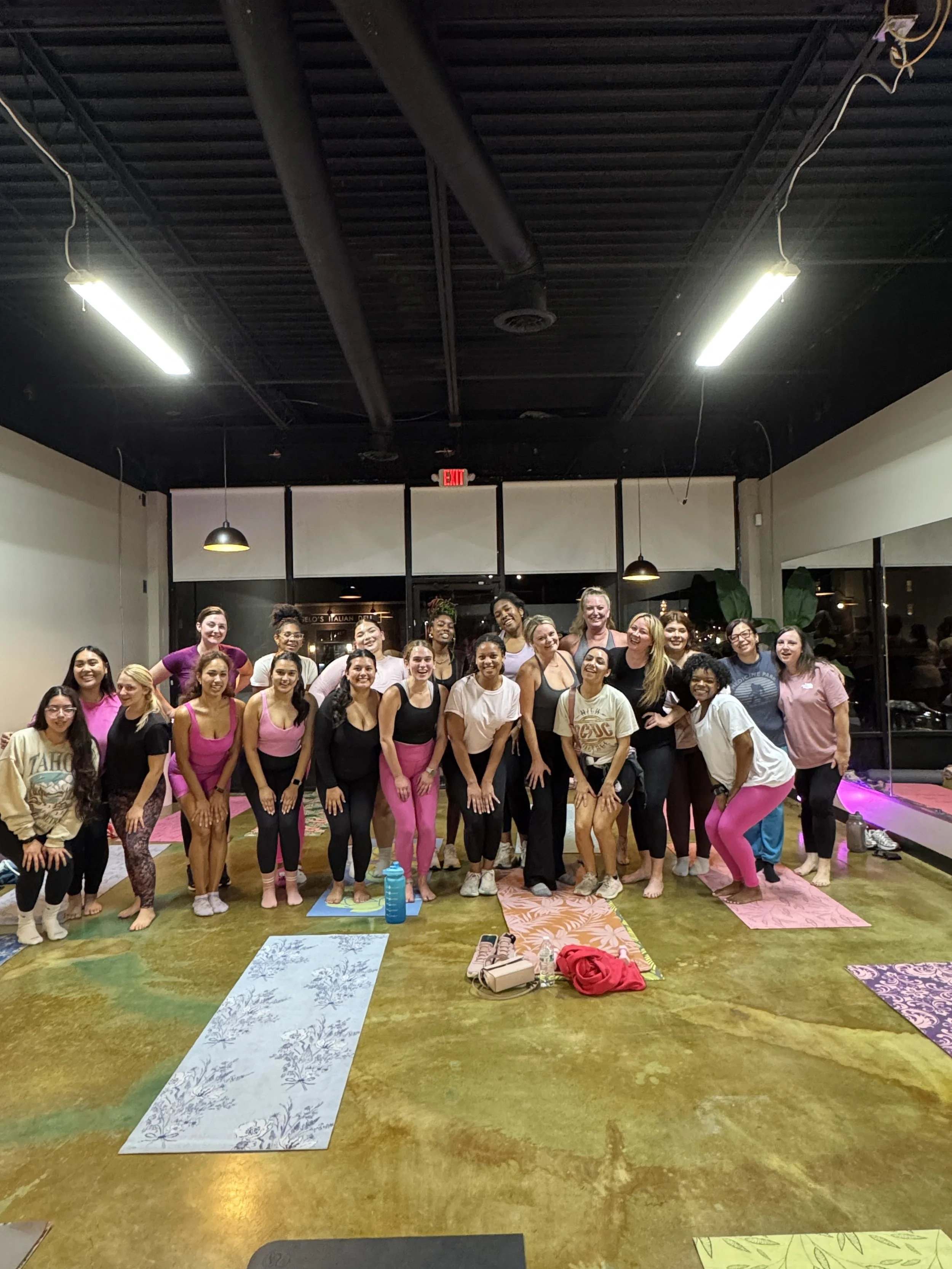 Group of women in yoga class, standing on yoga mats inside a studio, smiling at the camera.