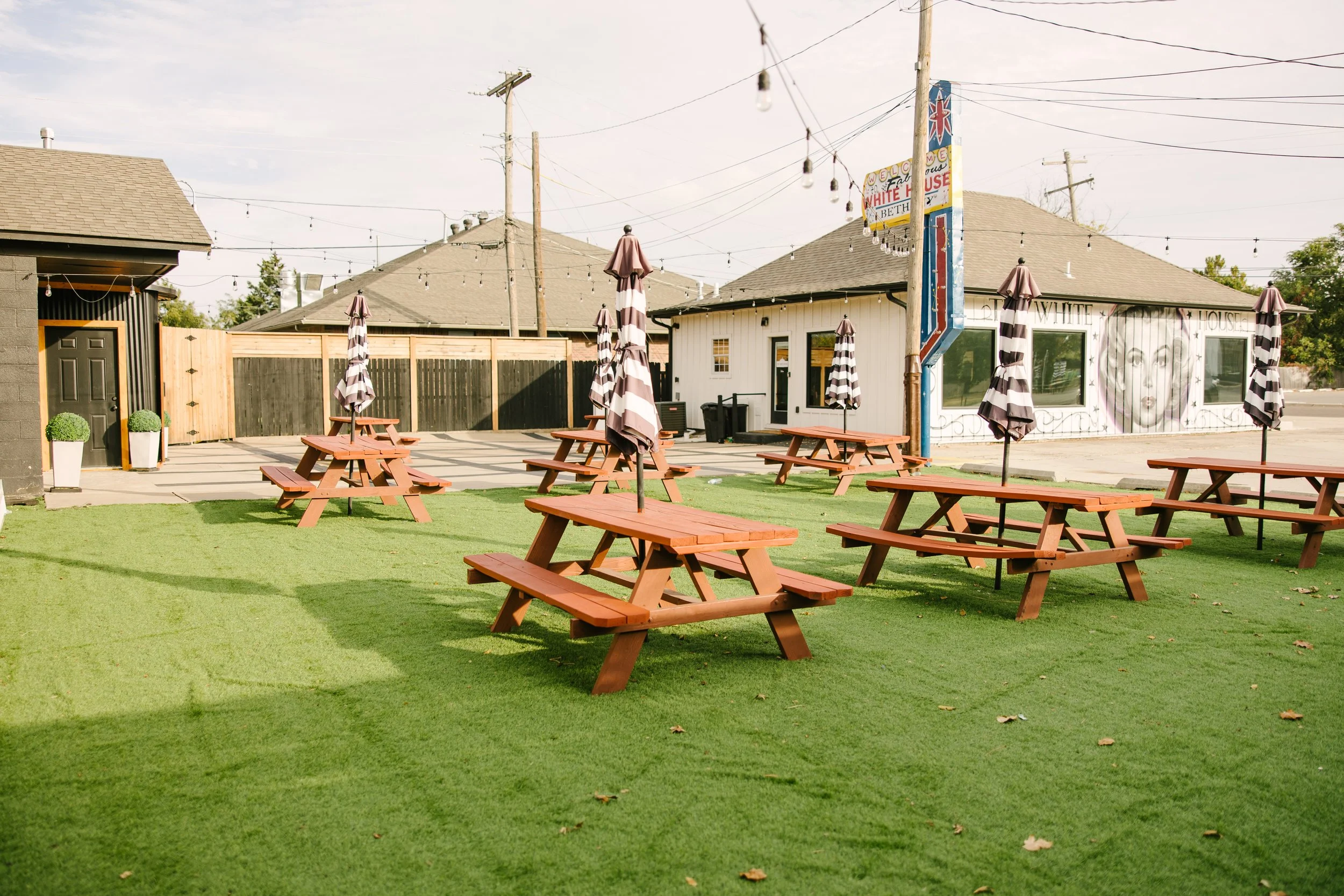 Outdoor patio with multiple wooden picnic tables with umbrellas, green artificial turf, a building with mural art, string lights overhead, and a fence in the background.