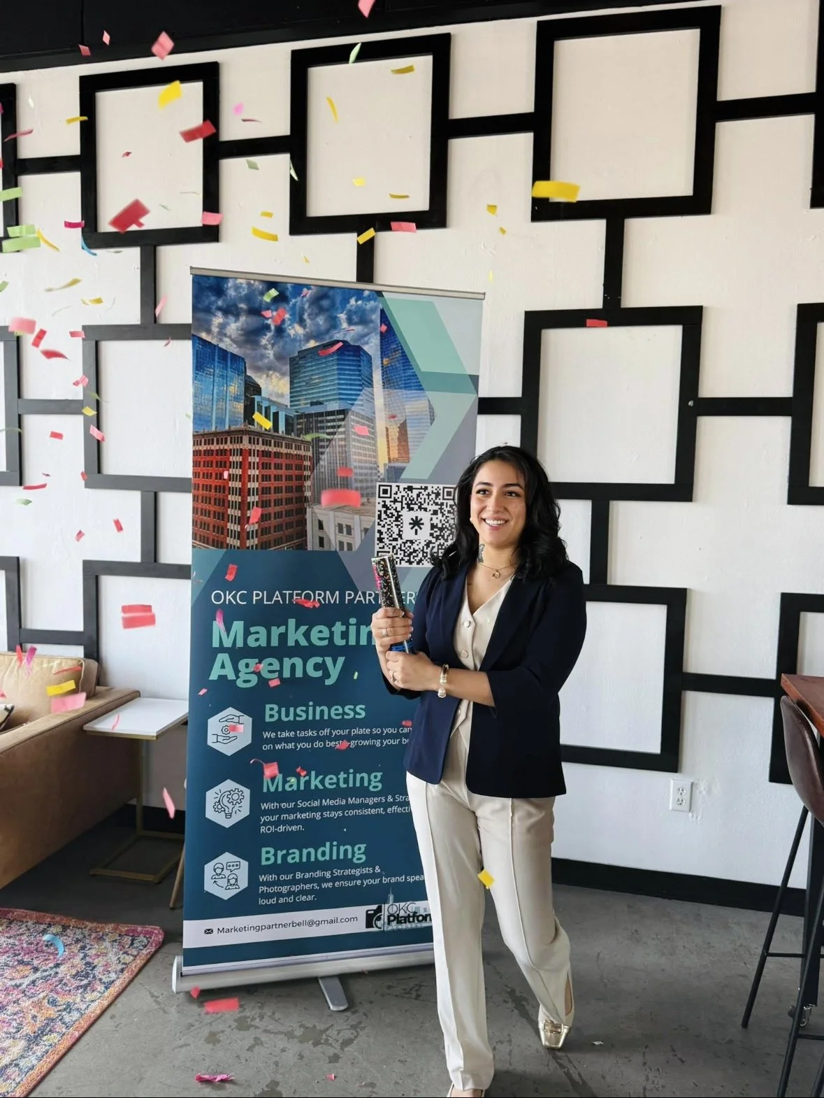 A woman in formal attire celebrating at a business event, standing next to a sign for an OKC marketing agency, with confetti falling around her.