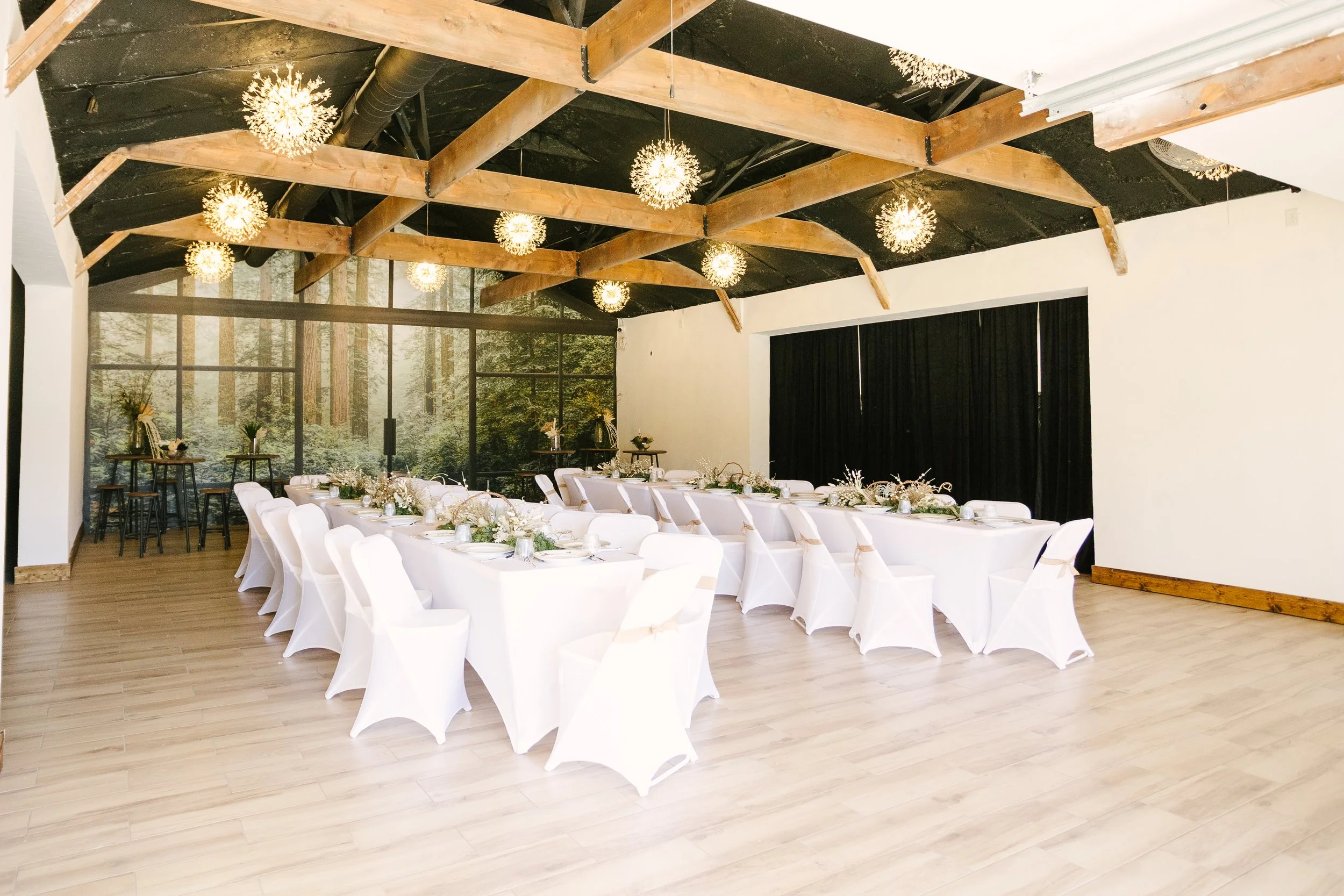 Wedding reception setup in a rustic indoor venue with long white-draped tables and chairs, floral centerpieces, and hanging spherical light fixtures on a dark wooden-beamed ceiling. Large windows show a forest outside.