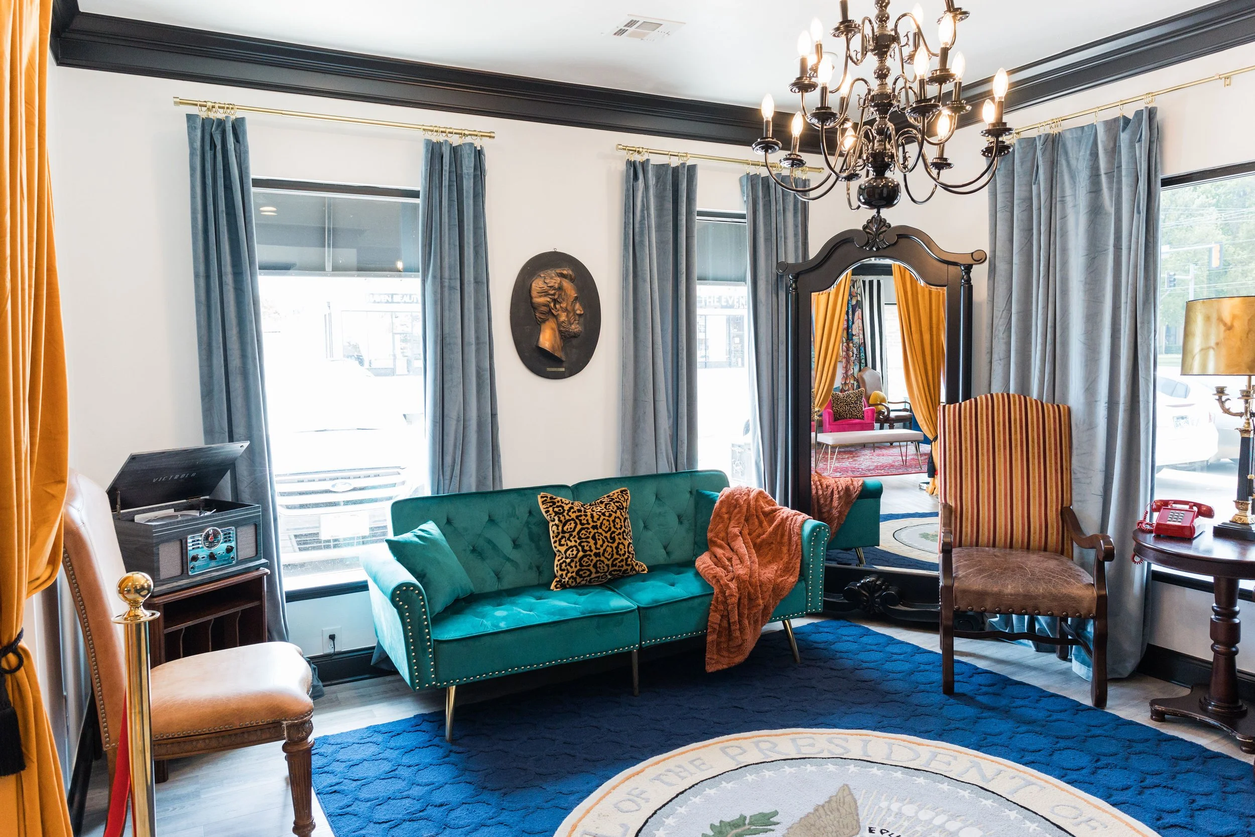 Living room with teal sofa, patterned pillow, orange throw blanket, vintage chairs, chandelier, and large windows with blue curtains.