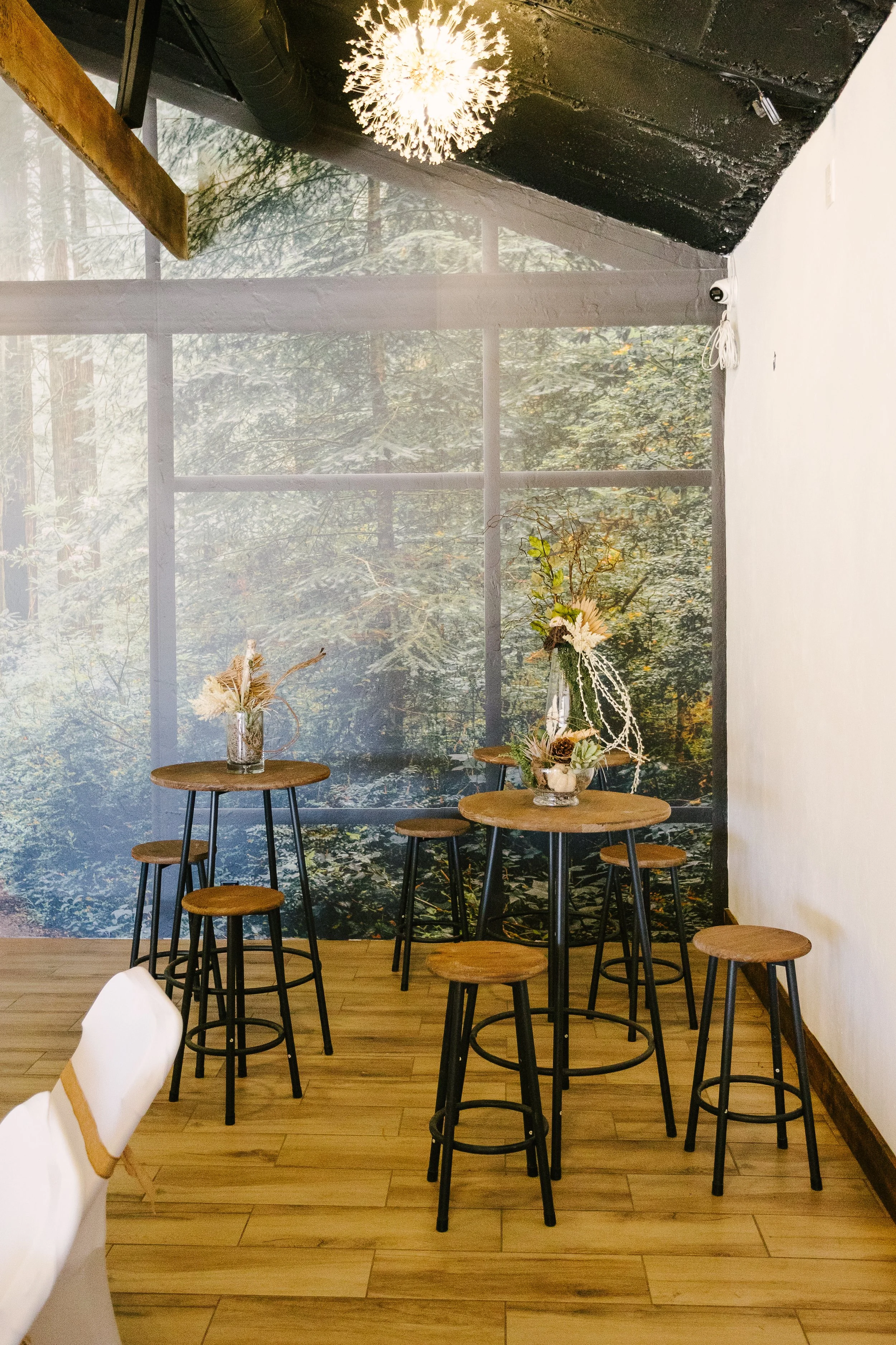 Interior of a cafe or restaurant with wooden tables and stools, decorated with floral arrangements, and a forest scene mural on the wall in the background.