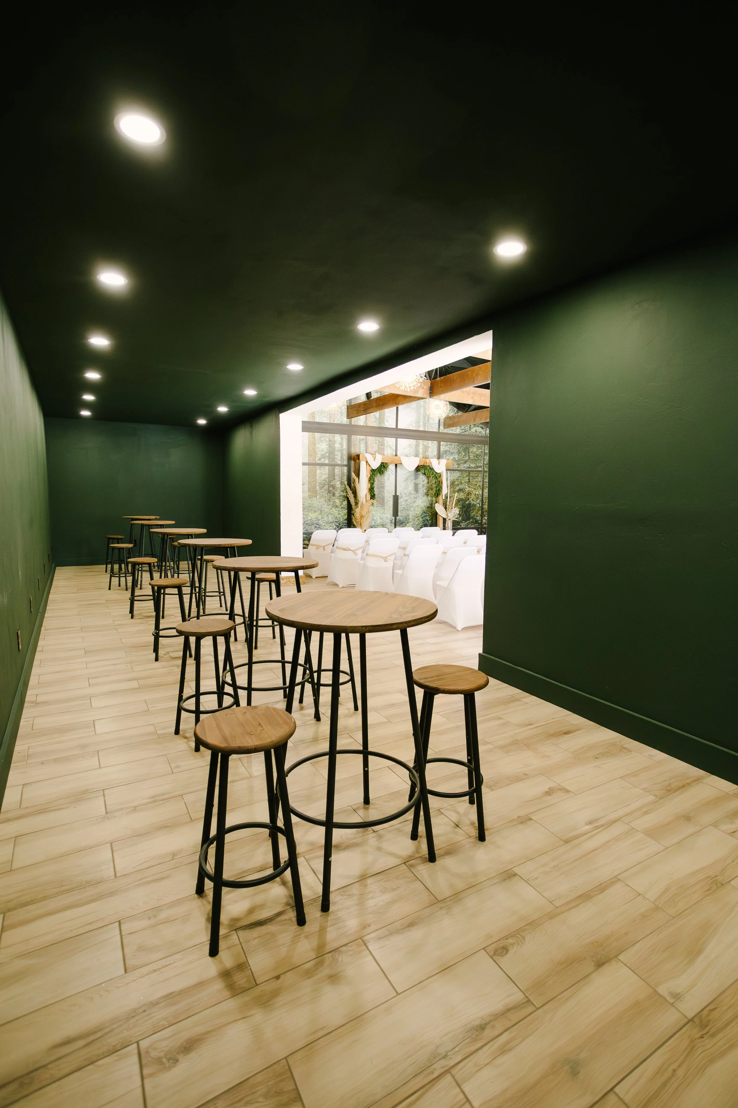 Empty green-painted room with wood floor, high tables and stools, leading to a decorated wedding ceremony area with white chairs and outdoors view.