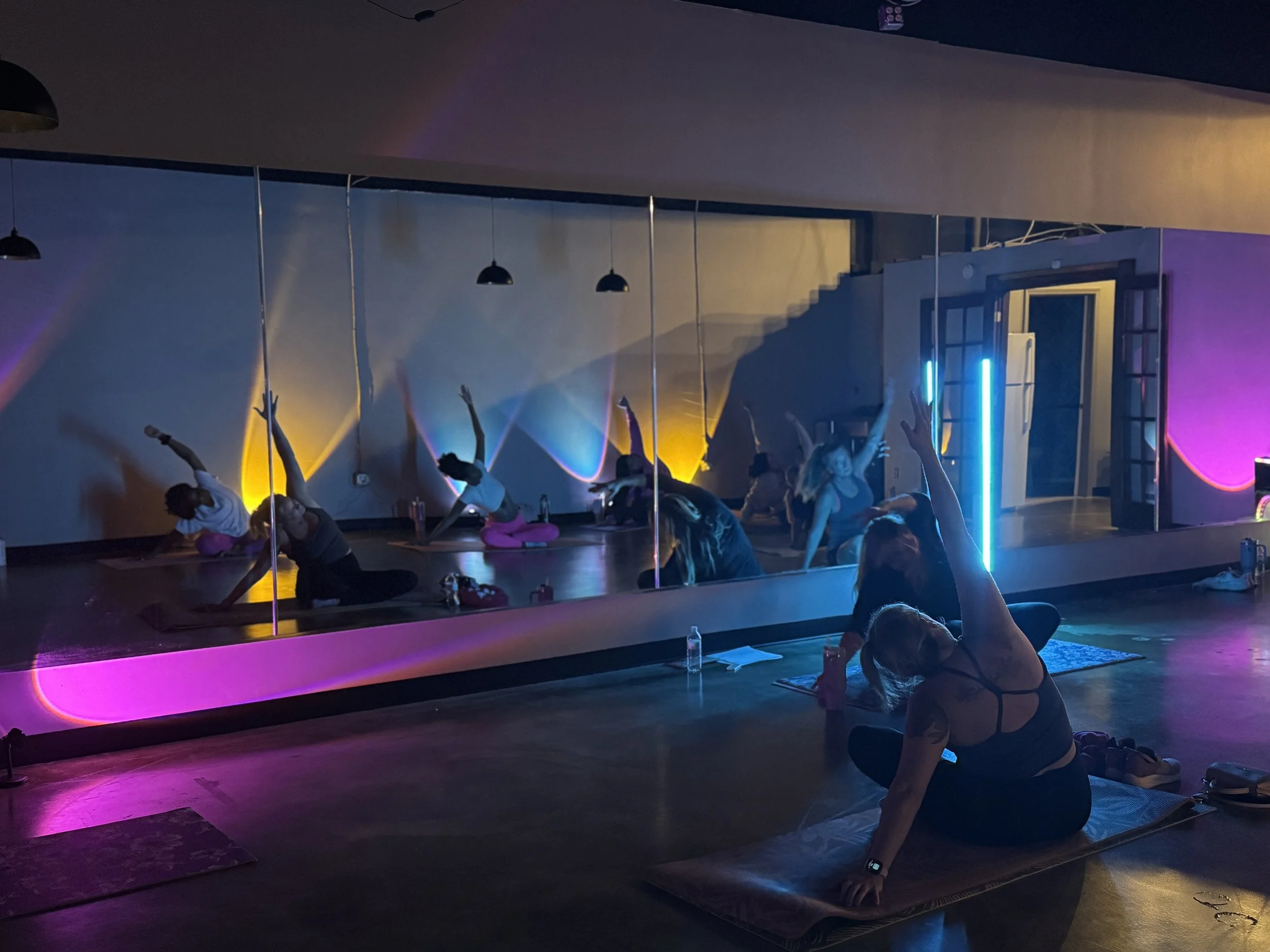A group of people participating in a yoga class in a dimly lit room with colorful lighting. Some are practicing poses near a mirror, while a woman in the foreground is sitting on a yoga mat, reaching upward with one arm.