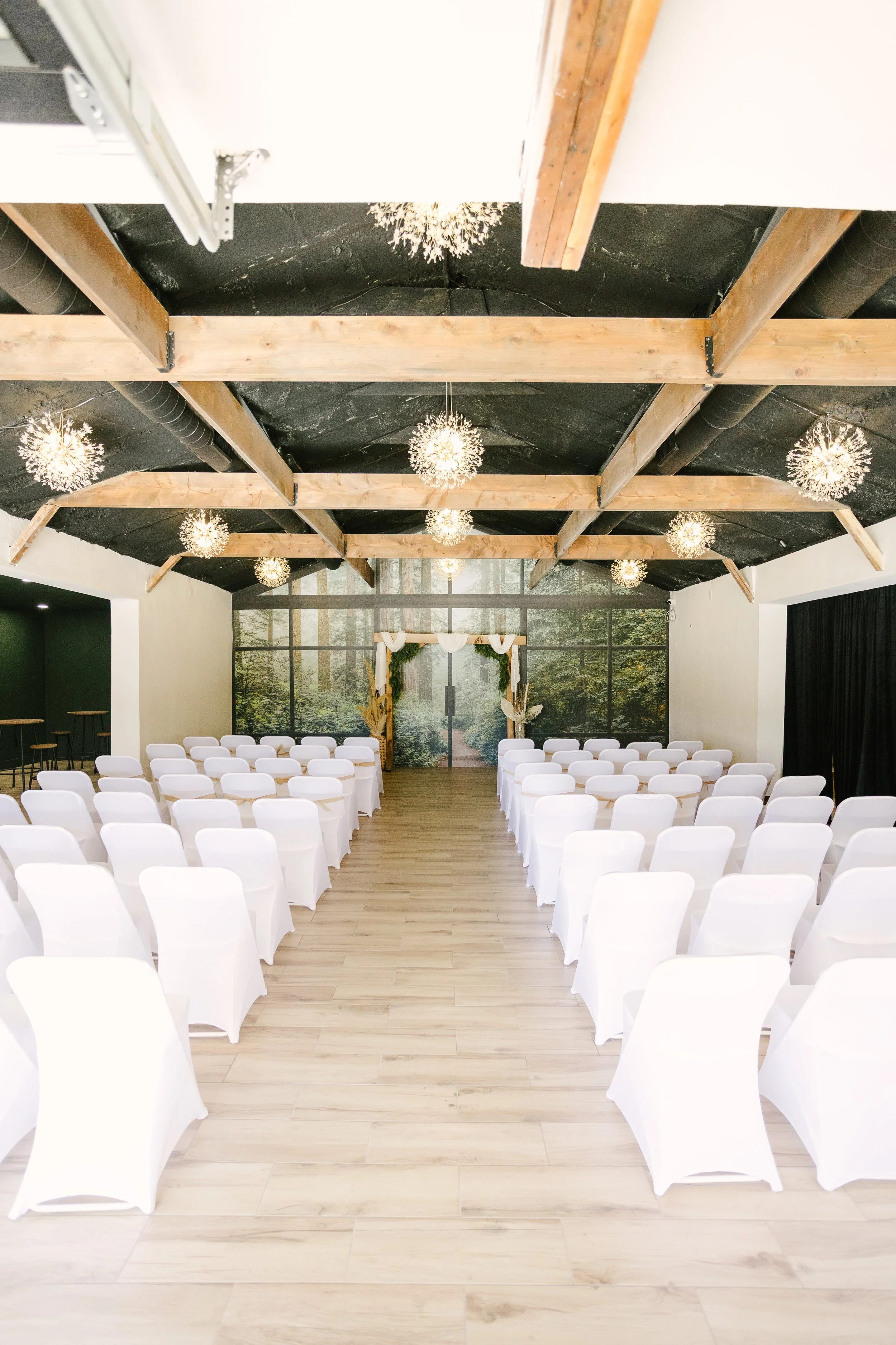 Indoor wedding ceremony setup with rows of white chairs, a floral arch at the front, and woodland-themed backdrop, ceiling with wooden beams and hanging spherical light fixtures.