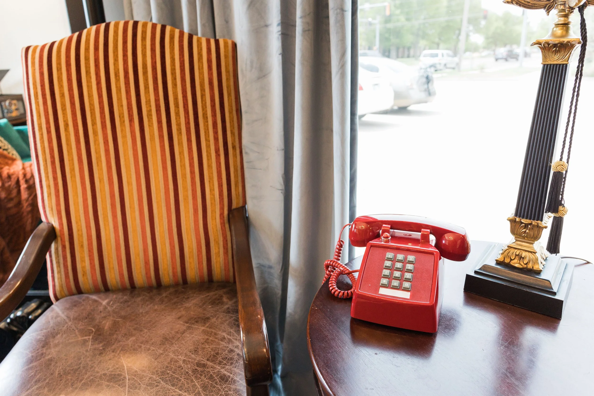 A red vintage rotary phone on a wooden table next to a lamp with black and gold decorative elements, with a striped upholstered chair nearby, and a window with curtains showing cars outside.