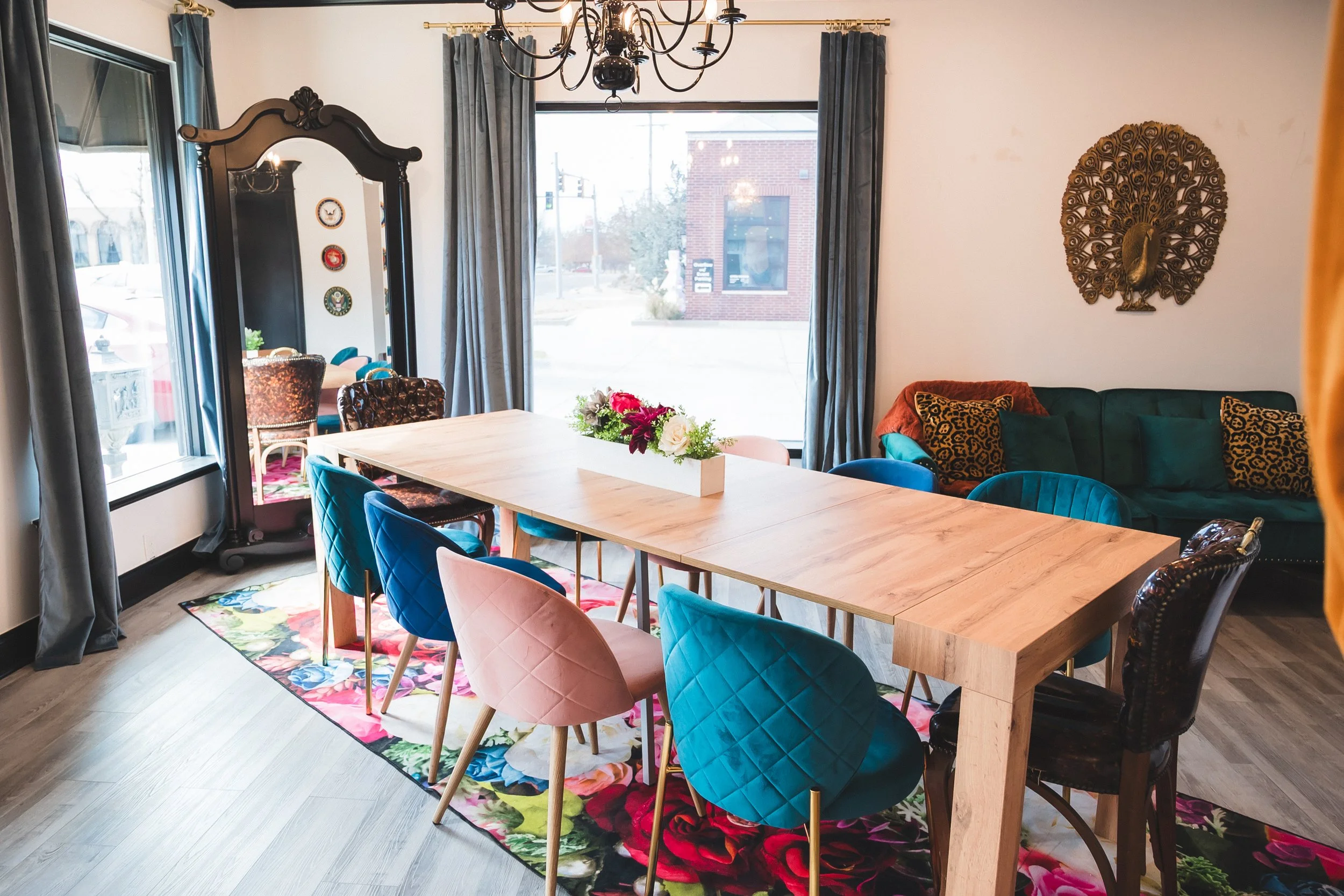A dining room with a long wooden table surrounded by colorful chairs, a floral centerpiece, a chalkboard, large windows with blue curtains, a green sofa with patterned pillows, and decorative wall art, illuminated by a chandelier.