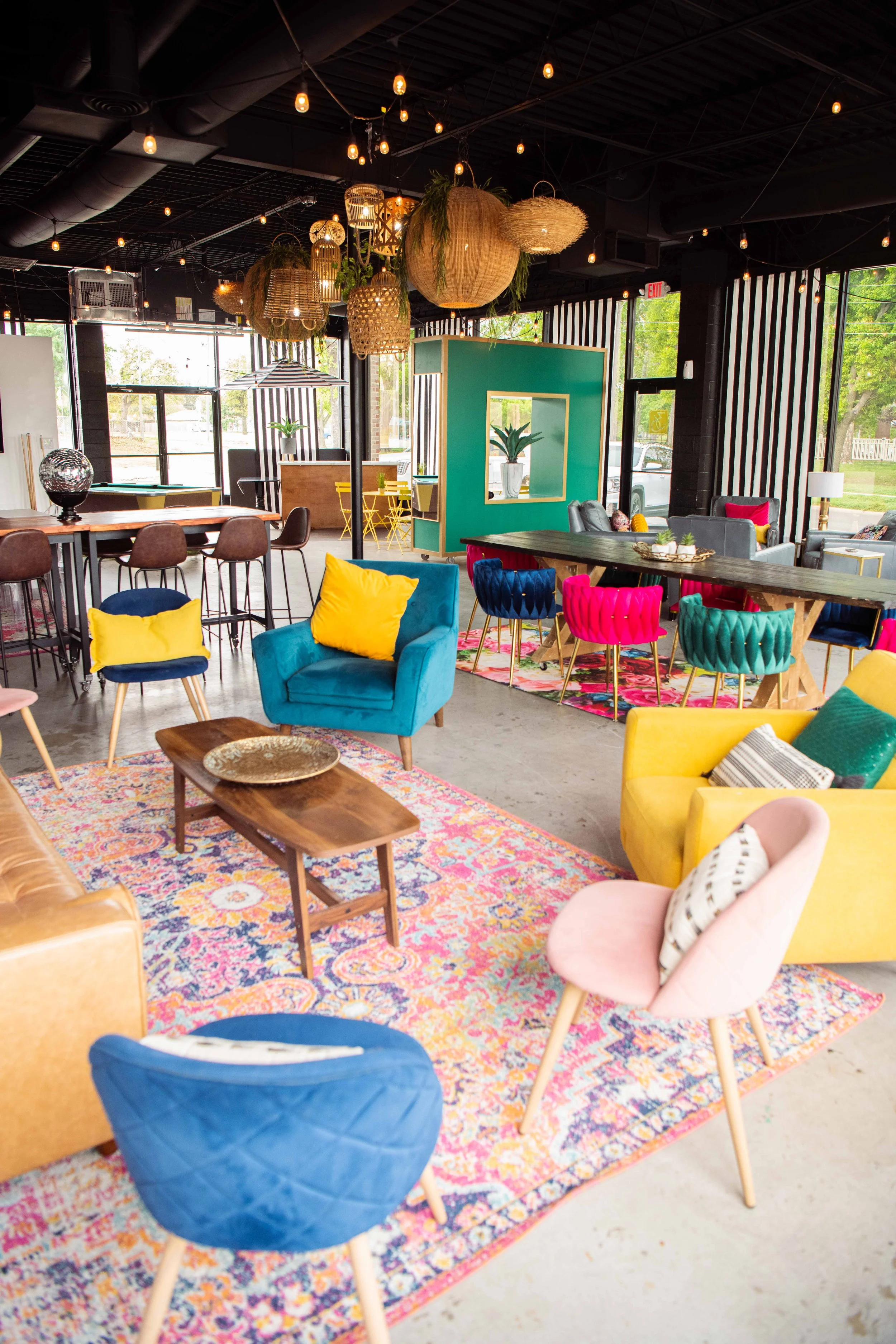 Interior of a colorful, modern lounge with various chairs, sofas, a patterned rug, hanging light fixtures, and decorative plants.