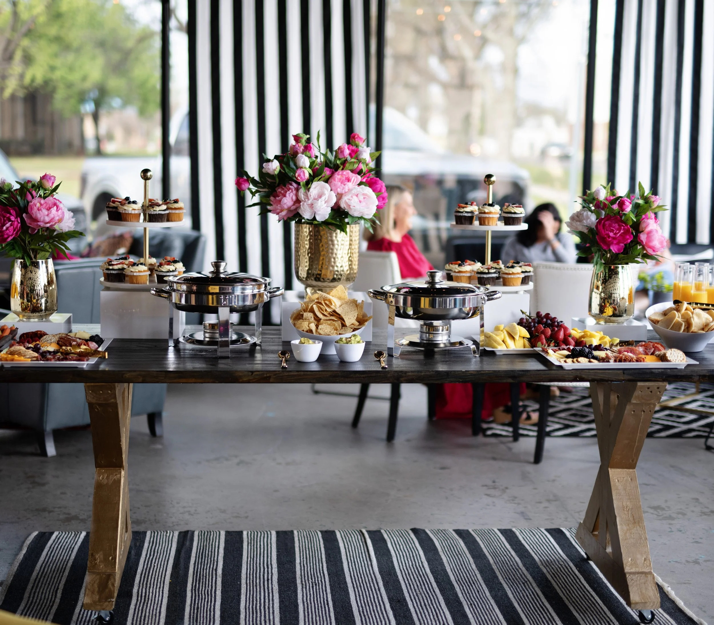 A buffet table with pink and white flowers in gold and white vases, serving dishes with cupcakes, chips, fruit, and other snacks, set in a bright room with large windows and people seated in the background.
