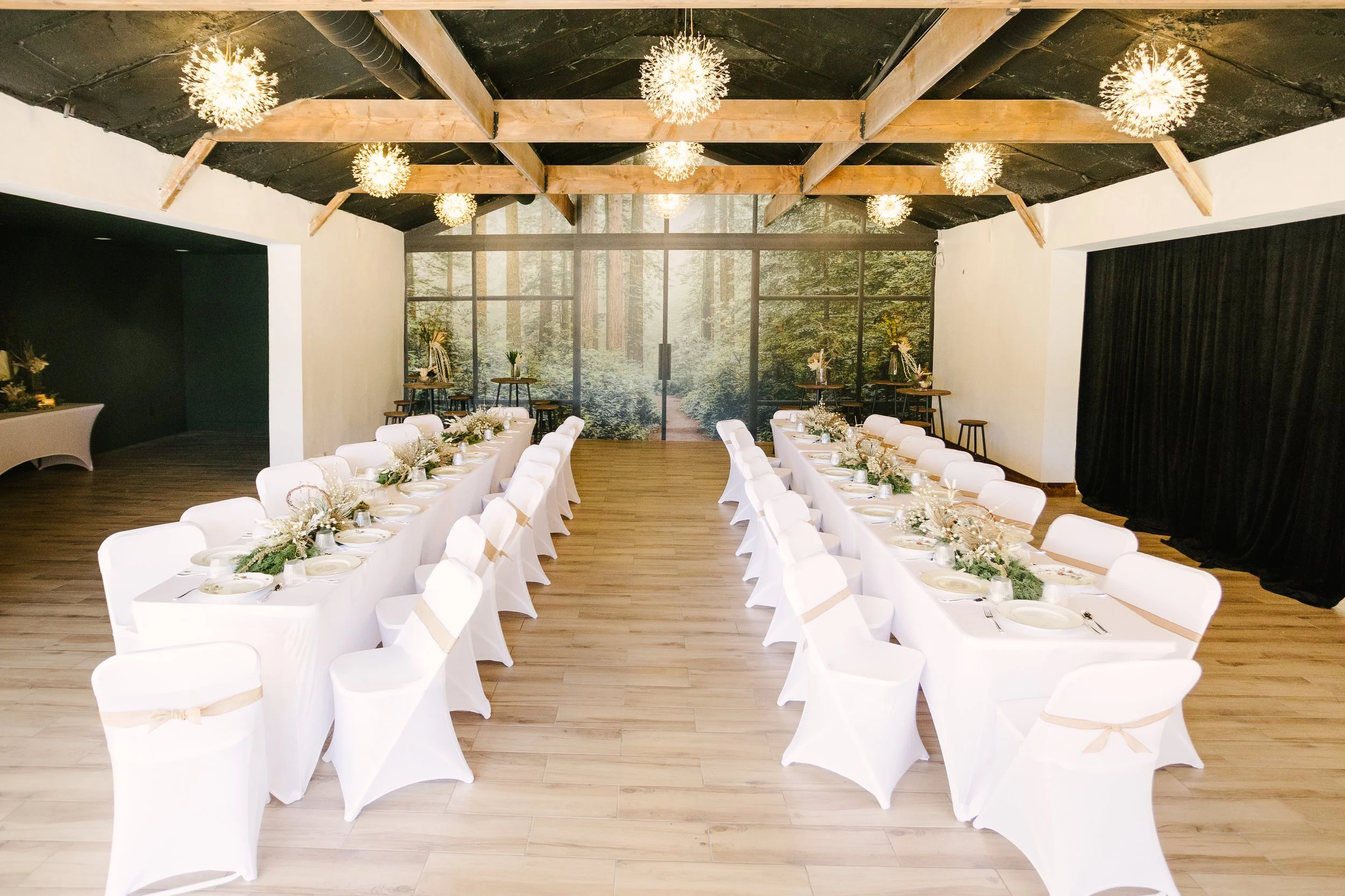 Wedding reception setup with long tables decorated with white tablecloths, floral centerpieces, and chairs covered in white fabric with beige sashes. The room has a wooden ceiling with light fixtures resembling dandelions and a forest mural on the ba