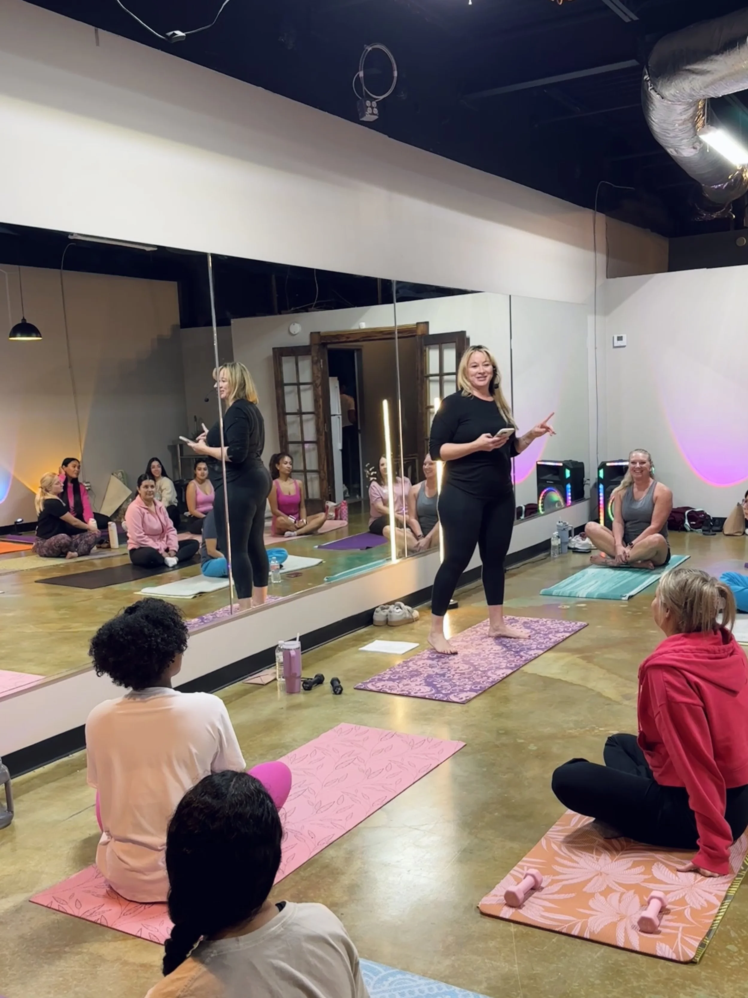Yoga class in progress with a woman instructor speaking to students in a studio with mirrors, yoga mats, and colorful lighting.