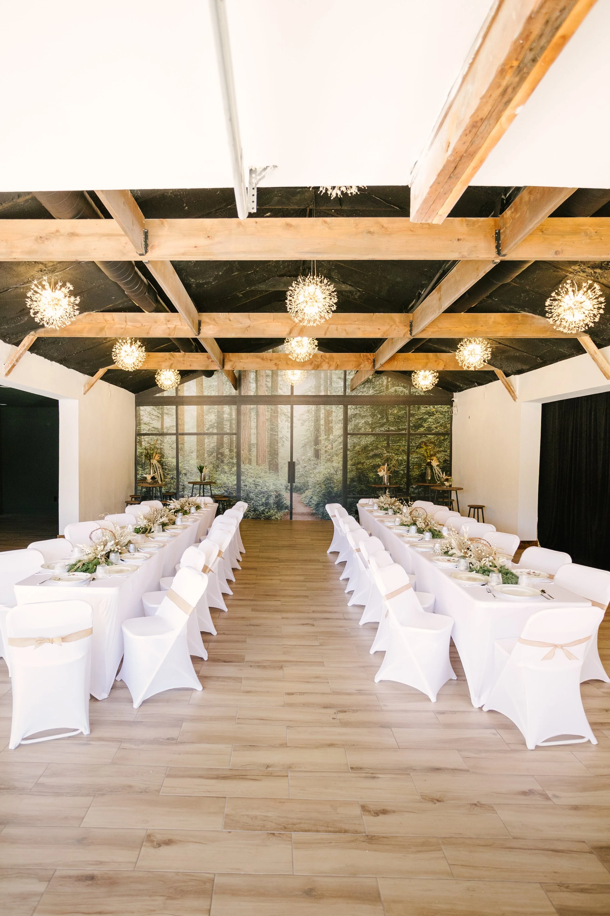 A decorated banquet hall with double tables arranged in a U-shape, white chair covers with beige ribbons, and table centerpieces of white flowers and greenery. The room has a high wooden-beamed ceiling with hanging spherical chandeliers and a large m