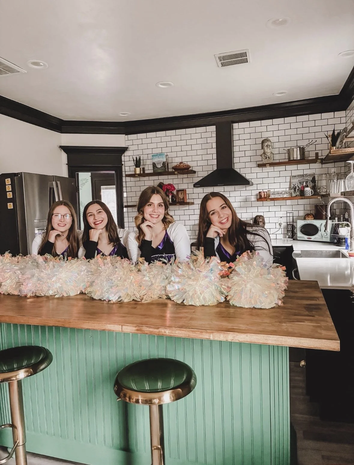 Four young women sitting at a kitchen counter, smiling, with colorful pom-poms in front of them, in a modern kitchen with white brick walls and black accents.