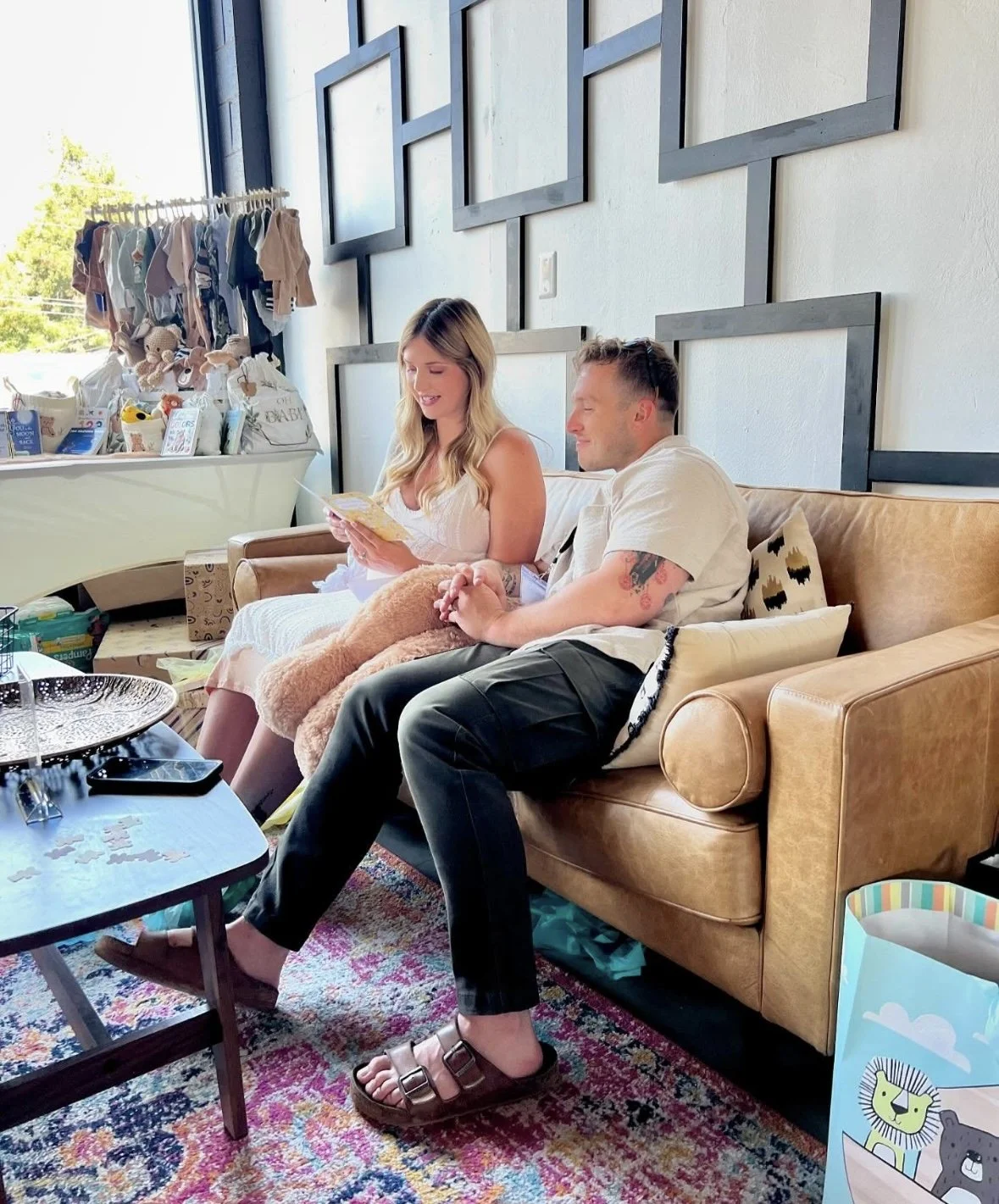 A woman and a man sitting on a tan leather couch in a bright living room, with the woman reading a card and the man holding her hand. Behind them, there is a display of baby clothes and gifts on a white surface, and holding a large baby teddy bear co