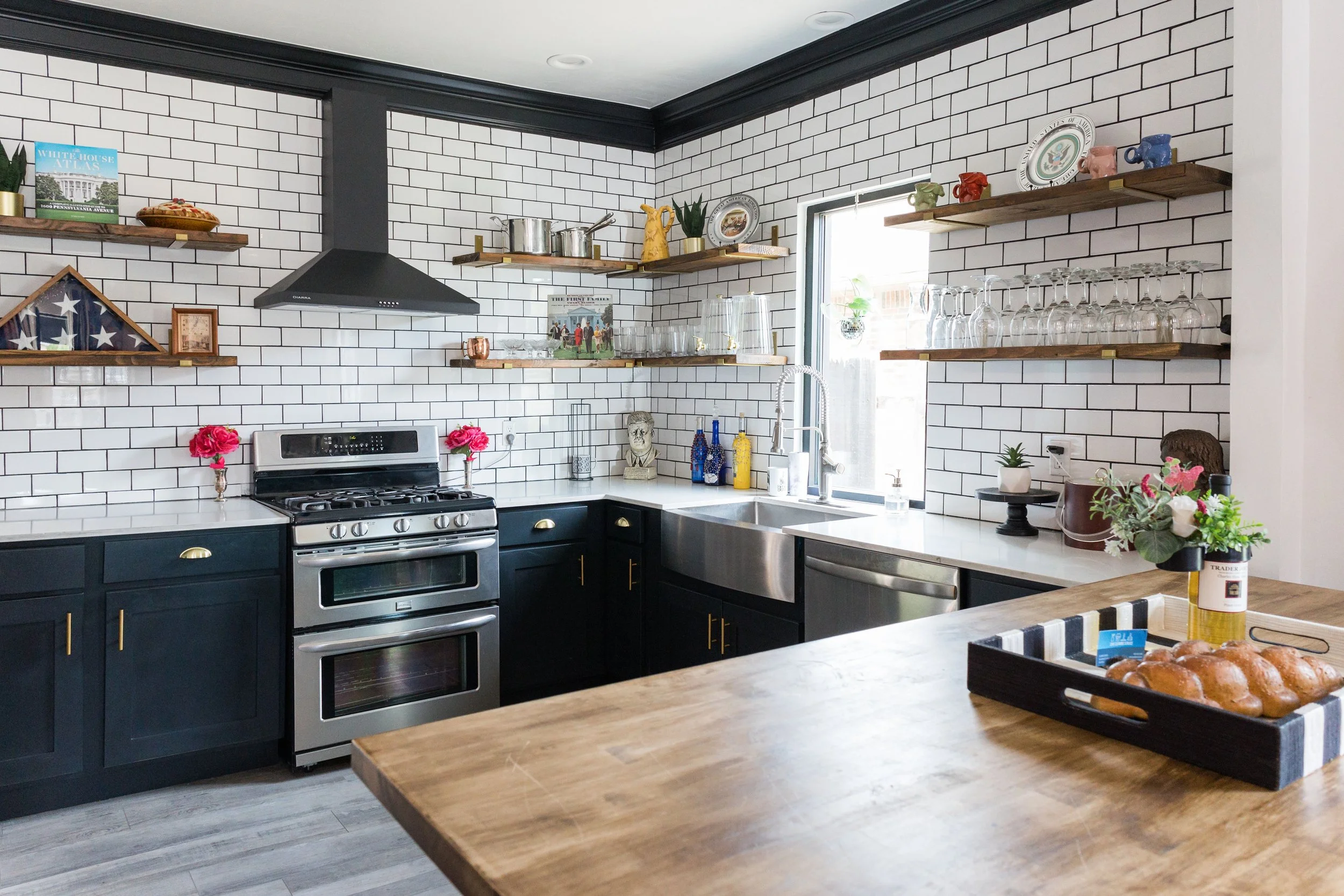Modern kitchen with white subway tile walls, black cabinets, stainless steel appliances, open wooden shelves with dishes and glassware, and a wooden island with flowers and a tray of bread.