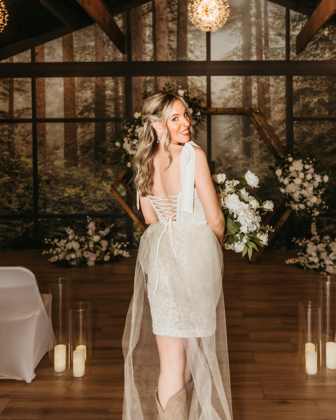 a woman in a white dress is getting bridal photos done. She is holding a white flower bouquet standing in front of a wedding arch and the background has a beautiful scenic view on the wall of having a forest like view where the view always stays gree