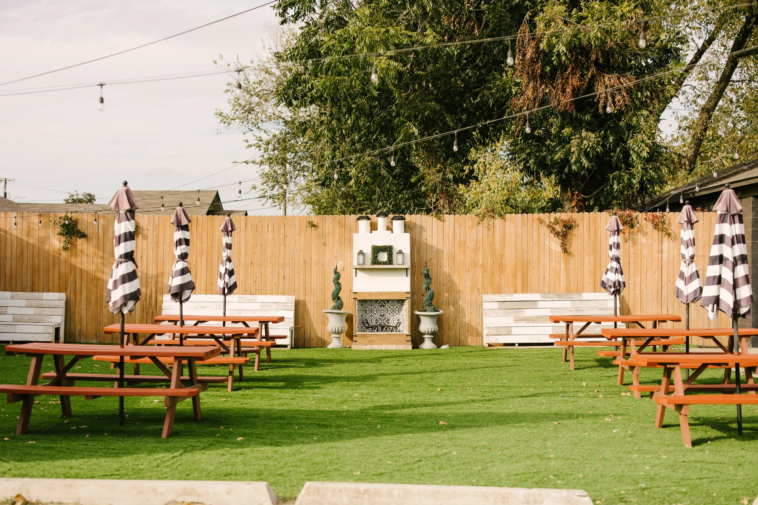 A backyard with green grass, tables with black and white striped umbrellas, a wooden fence, and white benches. Decorative plants and a white fireplace with candles and lanterns are arranged against the fence. String lights are hung overhead.