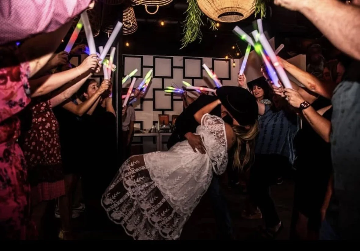 Couple kissing while surrounded by friends holding glow sticks during a celebration at a party or wedding reception.