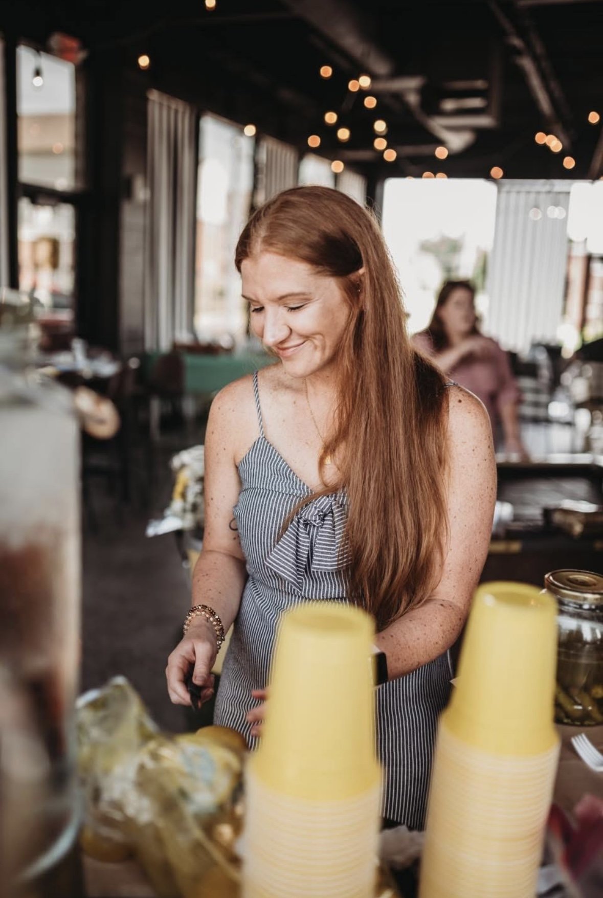 Smiling woman with long red hair and freckles, wearing a striped tank top with a bow, standing behind a counter with yellow disposable cups in a restaurant or cafe with string lights and large windows.
