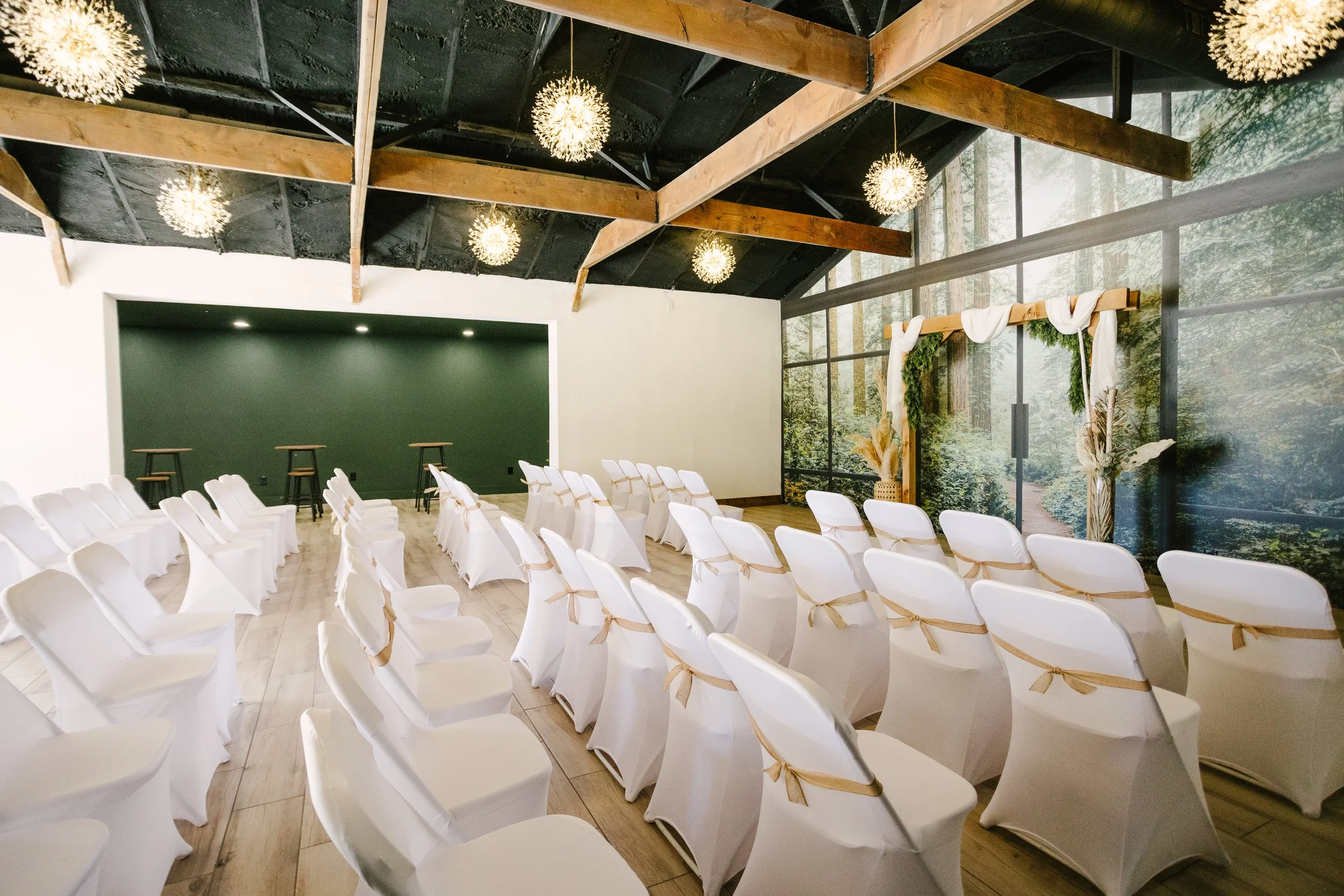 A decorated indoor wedding ceremony space with white chairs tied with beige ribbons, facing a wooden arch draped with white fabric and greenery. The room has a large mural of a forest scene on one wall, wooden beams, and hanging spherical light fixtu