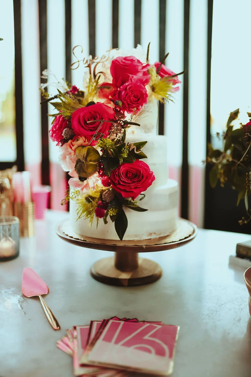 A white wedding cake decorated with pink roses and green foliage on a wooden cake stand, with a pink spatula and pink numbered cards on the table, and black slatted windows in the background.