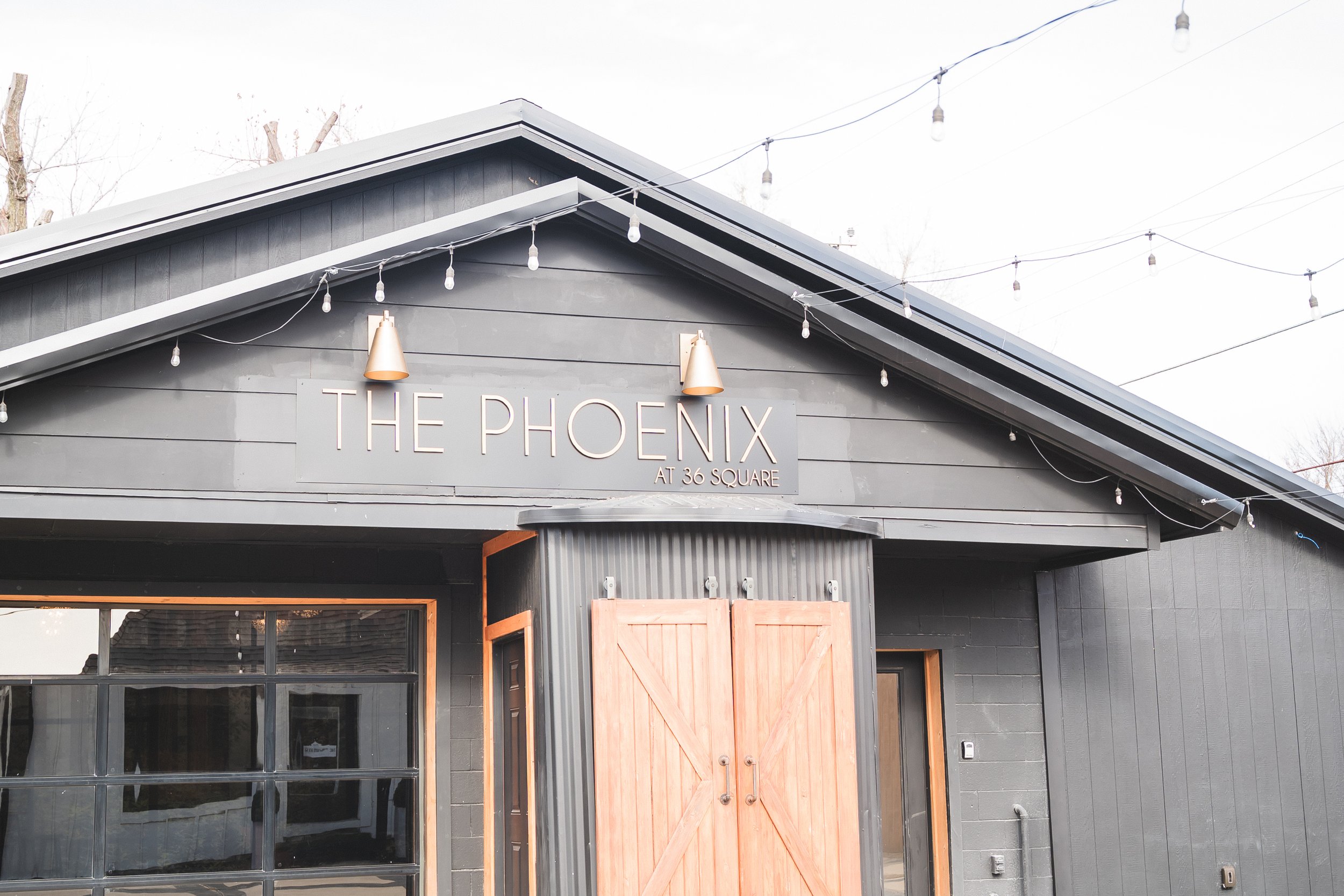 Exterior of a building named 'The Phoenix at 36 Square' with string lights hanging above and a large wooden door.