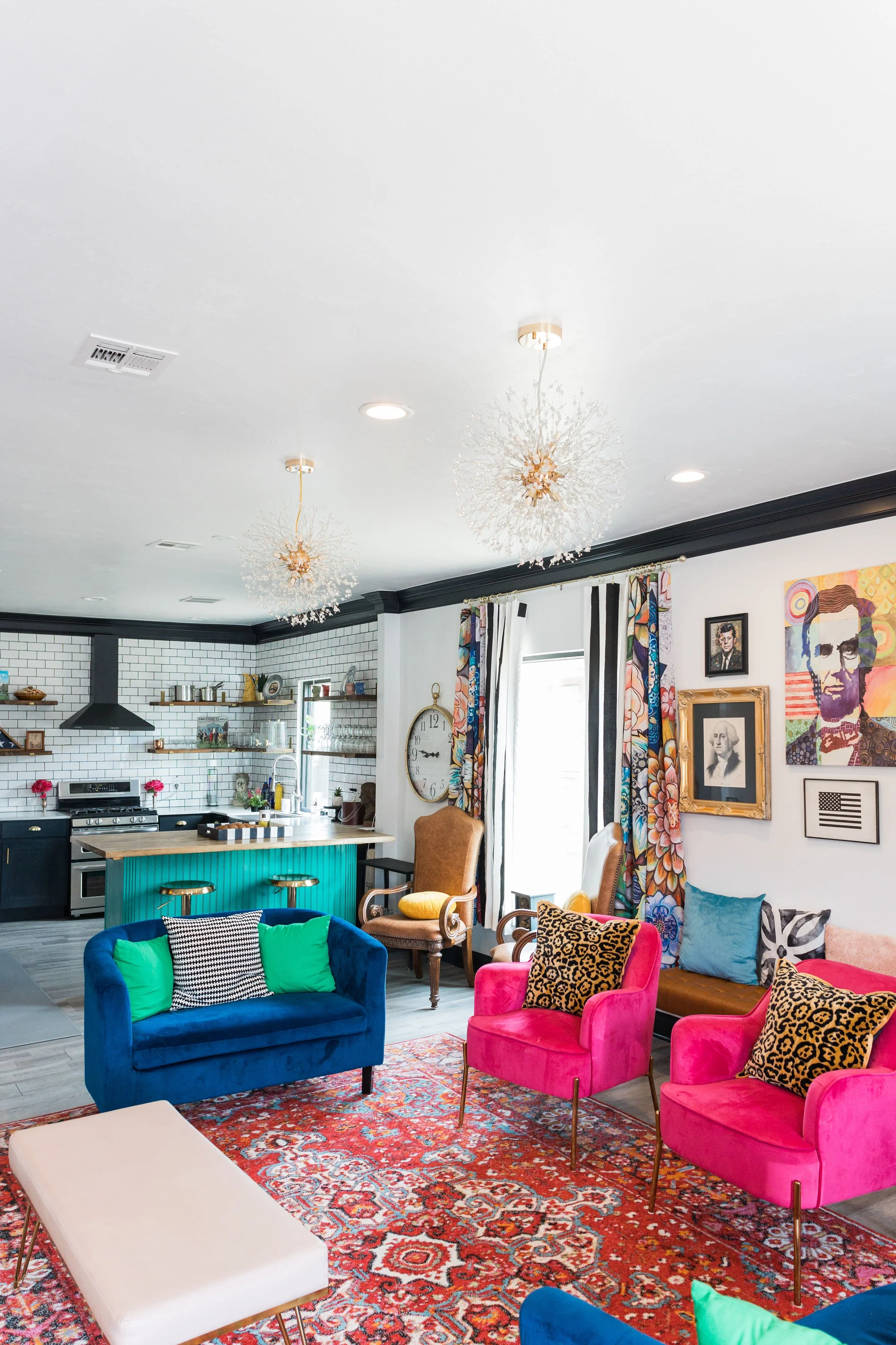 Colorful living room with pink and blue chairs, patterned cushions, a patterned rug, a kitchen area with open shelves, white subway tile backsplash, and black hardware, and framed portraits on the wall.