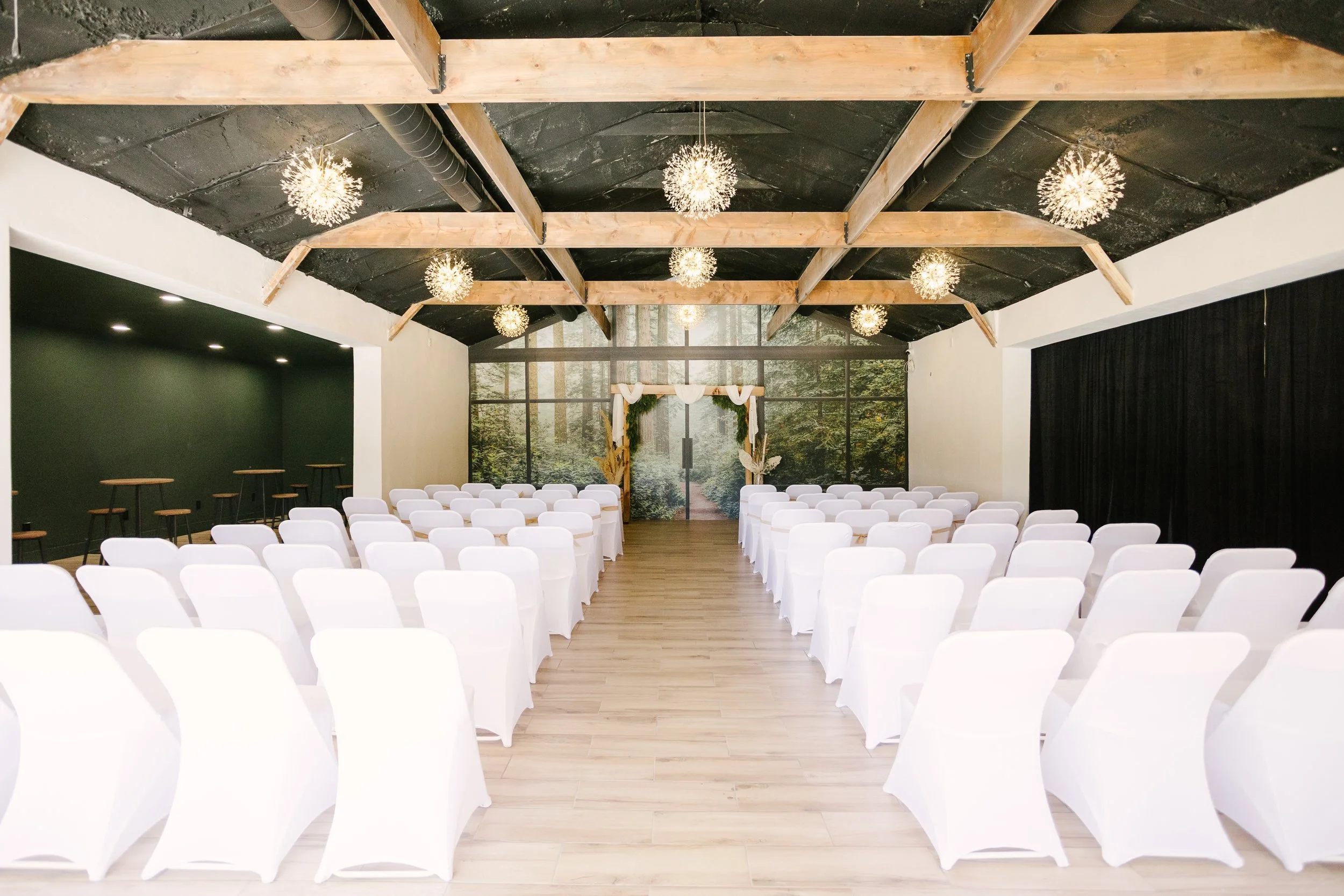 Indoor wedding ceremony setup with rows of white chairs facing an arch decorated with white fabric and greenery, large wall mural of a forest scene, black curtains on side, and wooden ceiling beams with hanging spherical light fixtures.